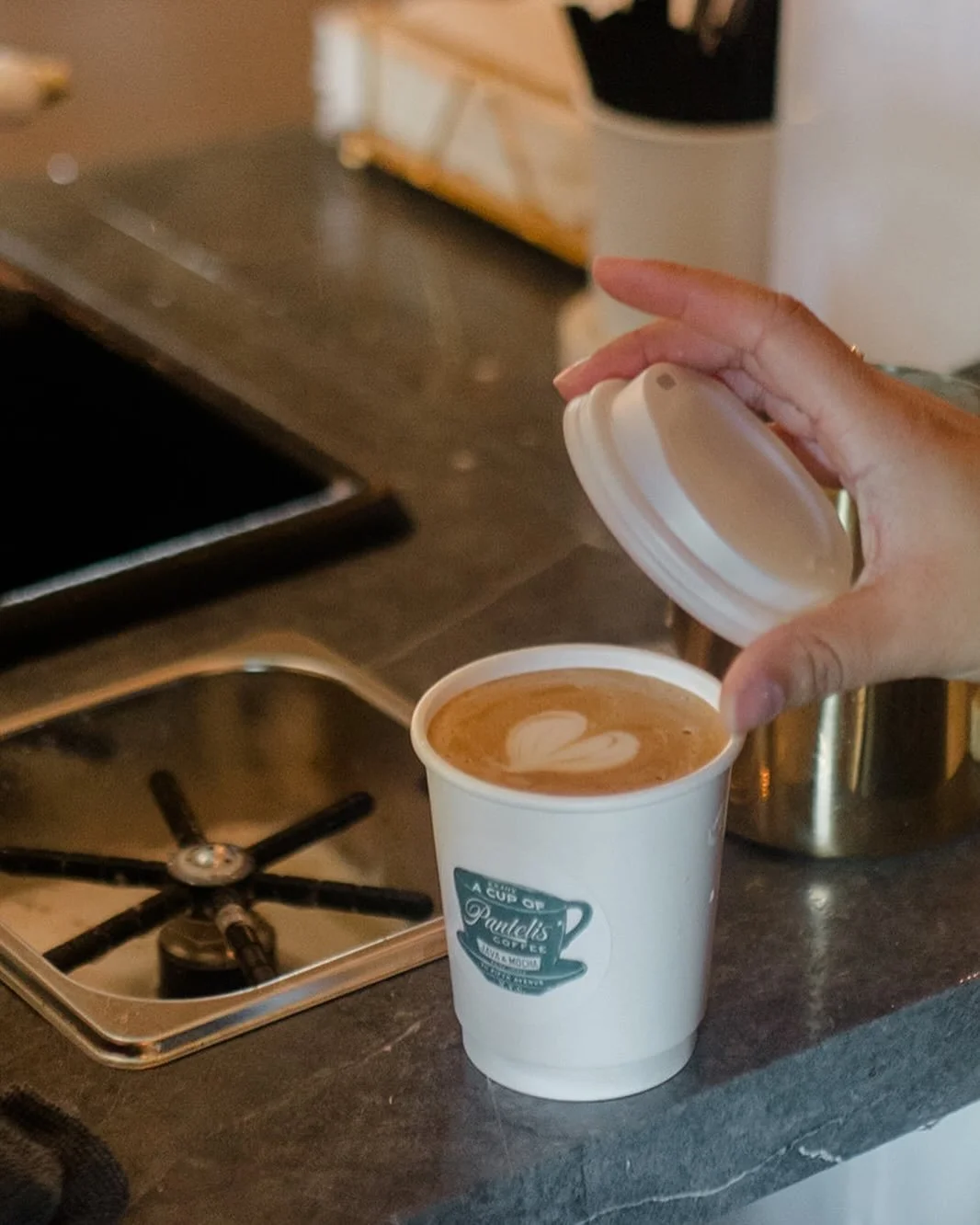 A hand holding the lid of a to-go coffee cup, revealing a latte with heart-shaped foam art, on a kitchen countertop near a stove.