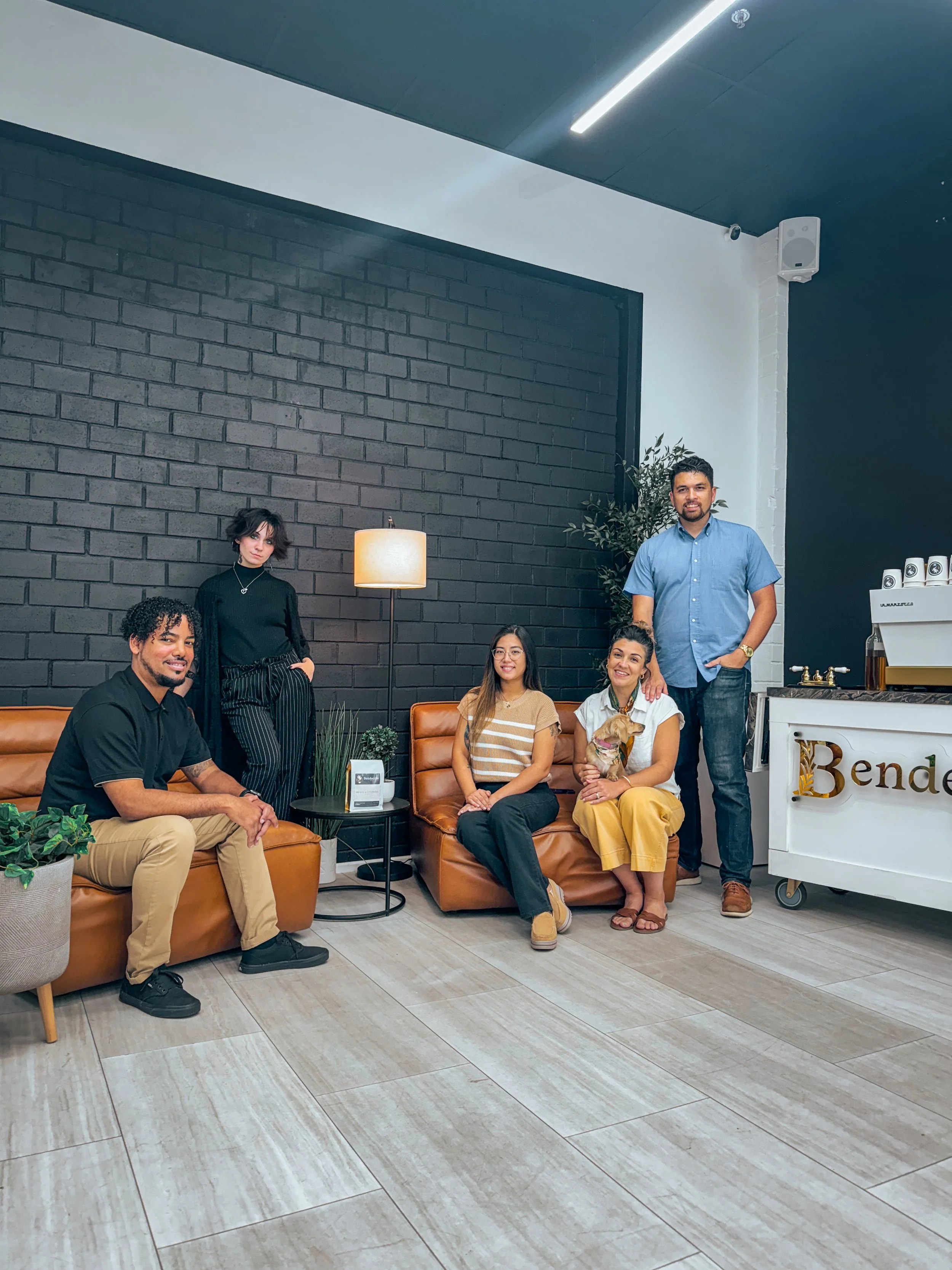 Group of five people, including a woman with a dog, sitting and standing in a modern lounge area with leather sofas, a black brick wall, and a sign that says 'Bendecido Coffee.'