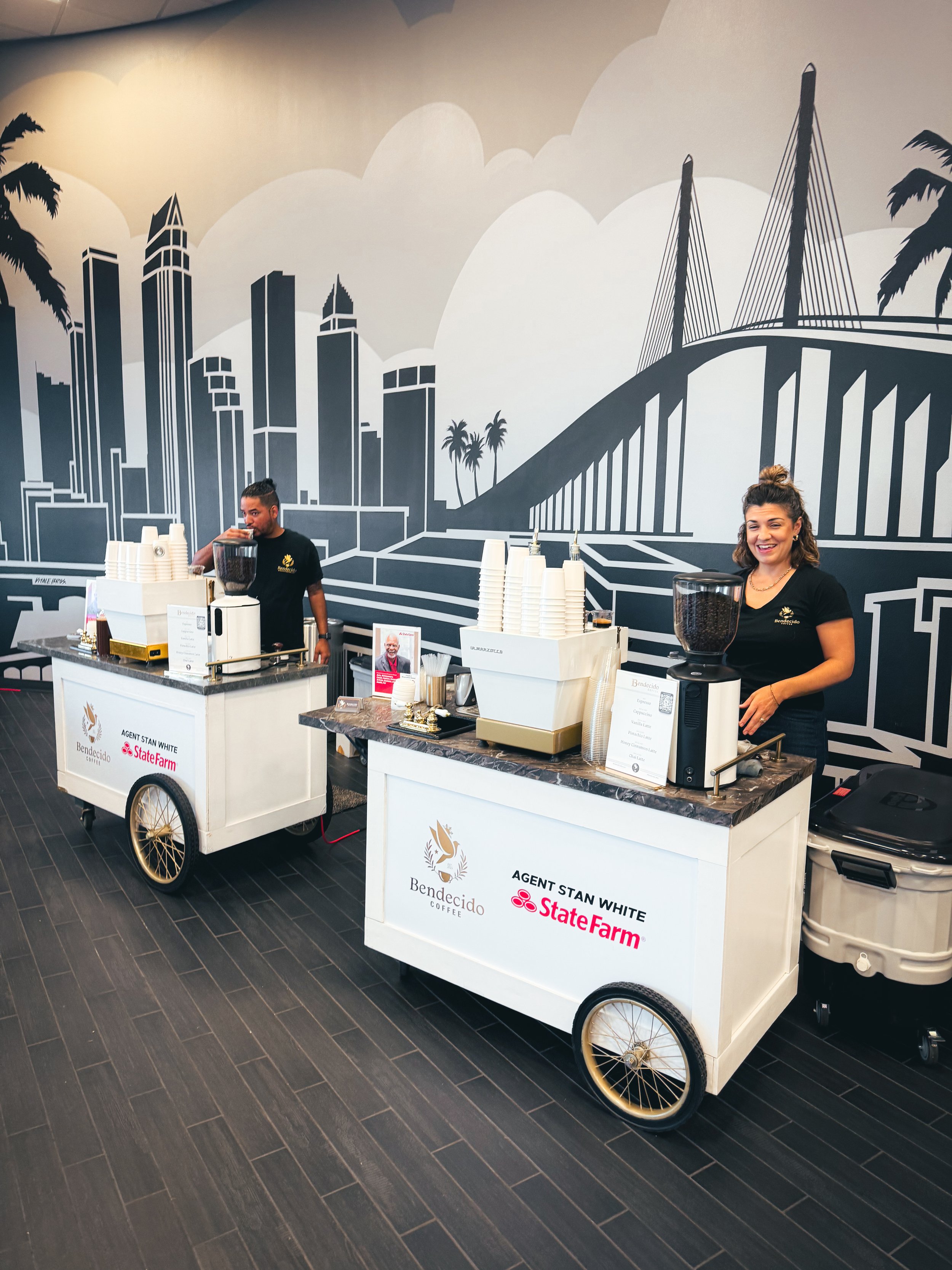 Two coffee carts with staff serving beverages in front of a city skyline mural, branded with Bendecido Coffee and State Farm logos.