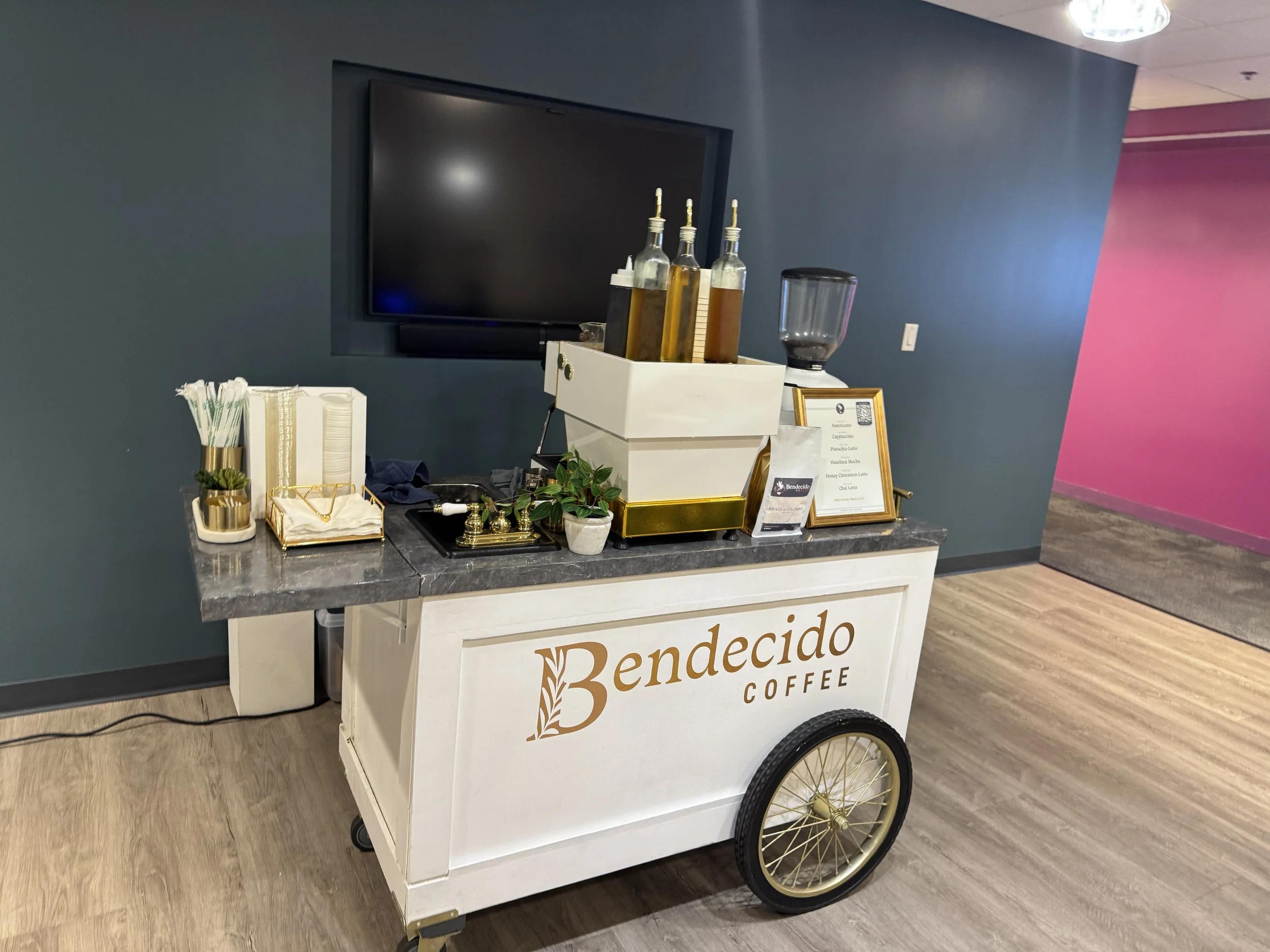 Coffee cart labeled Benedico Coffee with bottles of flavored syrup, cups, napkins, and a small plant, set against a dark blue wall with a television behind in a Tampa office.