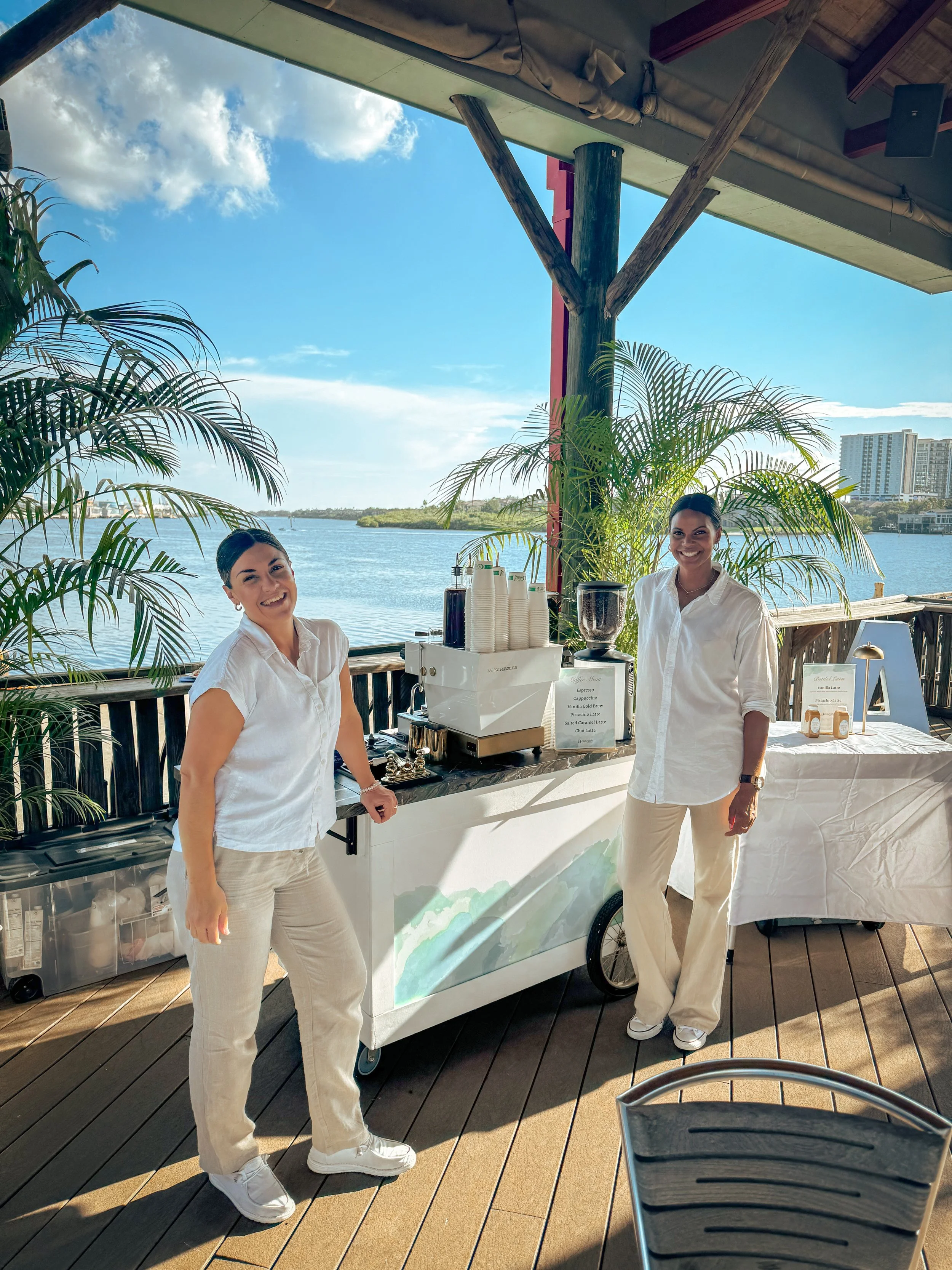 Two women in white shirts and light-colored pants stand next to a coffee station on a deck overlooking water, with palms and buildings in the background, smiling at the camera.