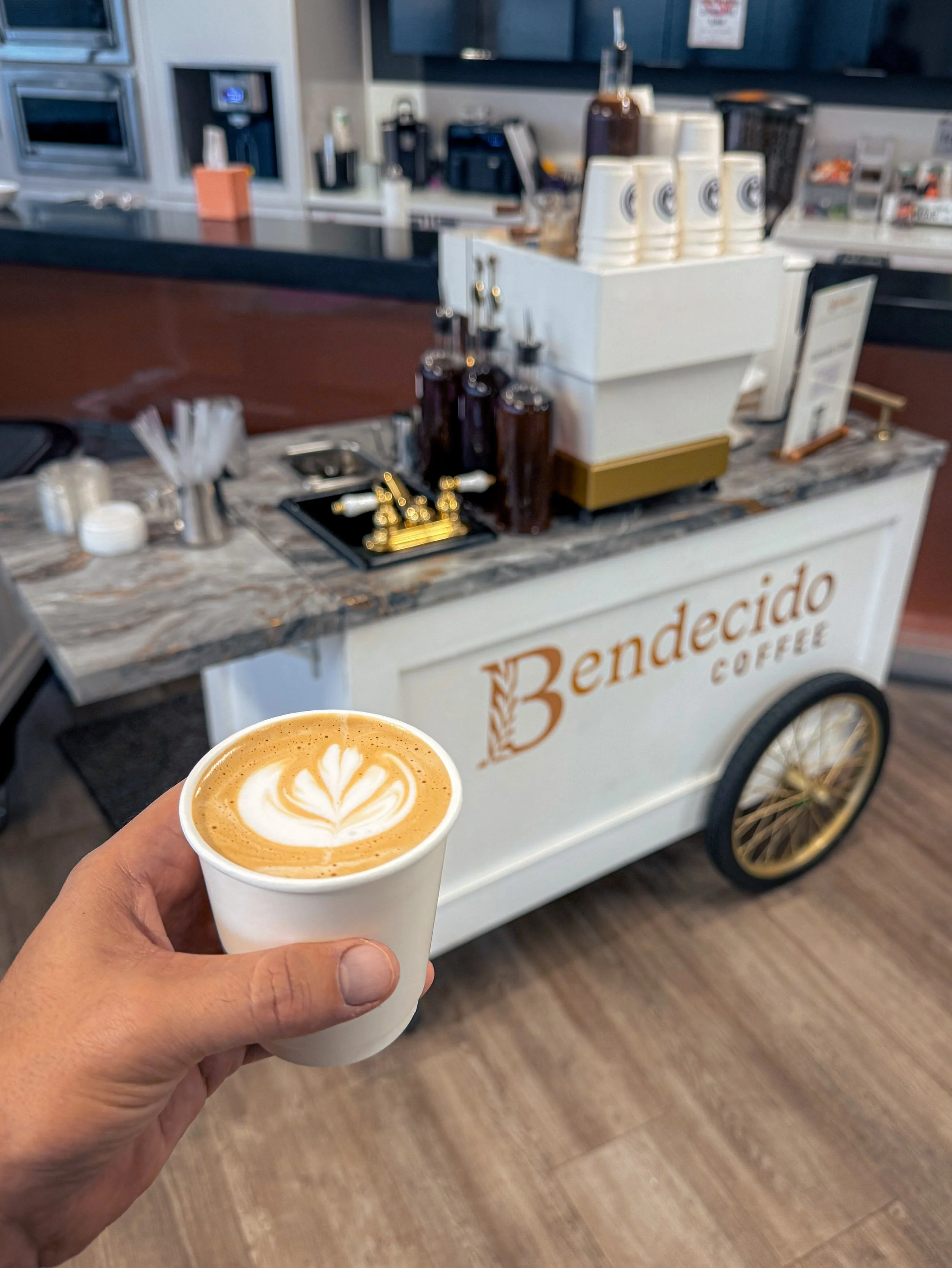 A person holding a cup of latte with latte art in front of a coffee cart labeled 'Benedeado Coffee' in a cafe.