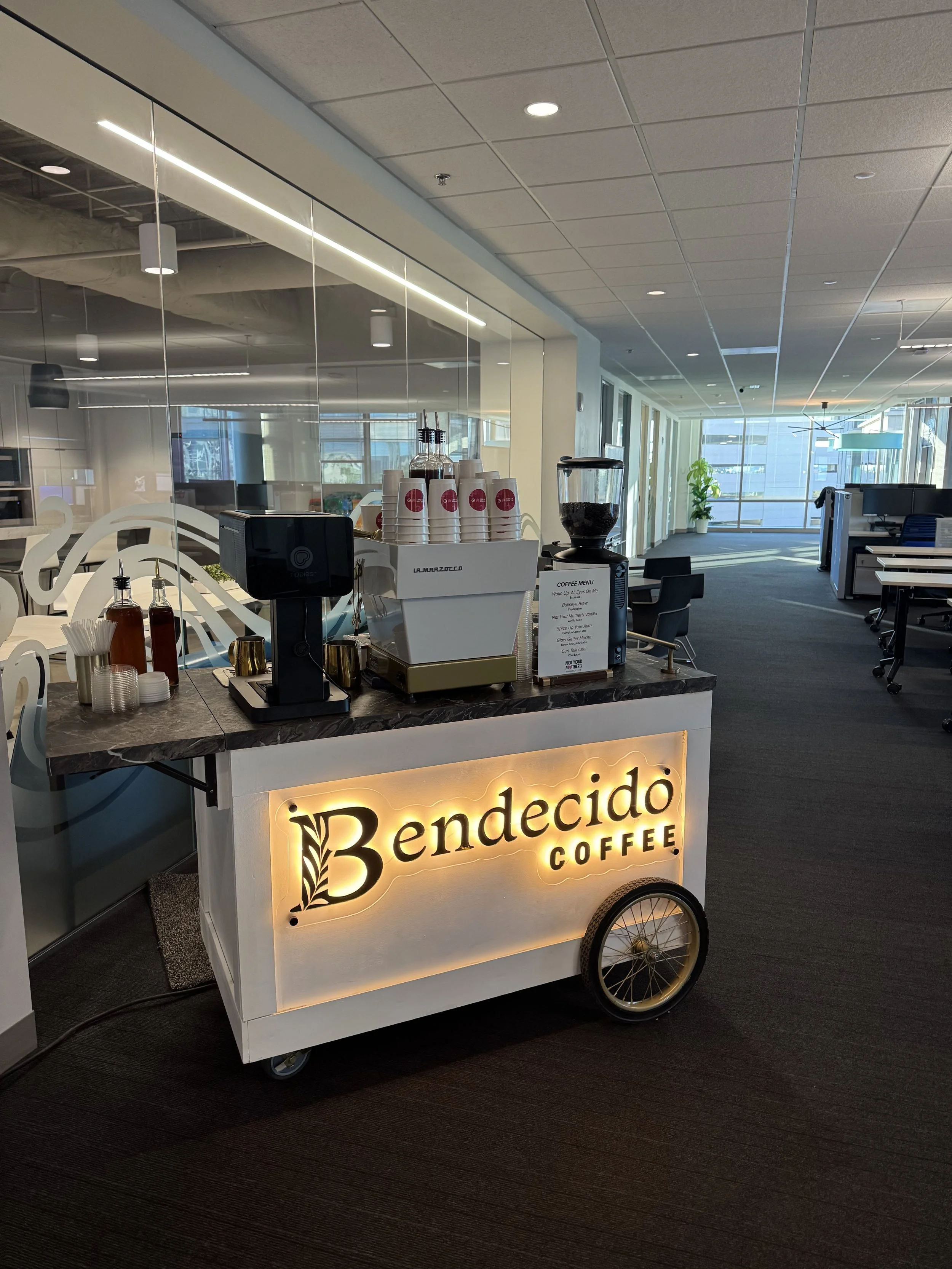 Mobile coffee cart with illuminated sign that reads 'Bendecido Coffee' located in an office environment with workstations and large windows.