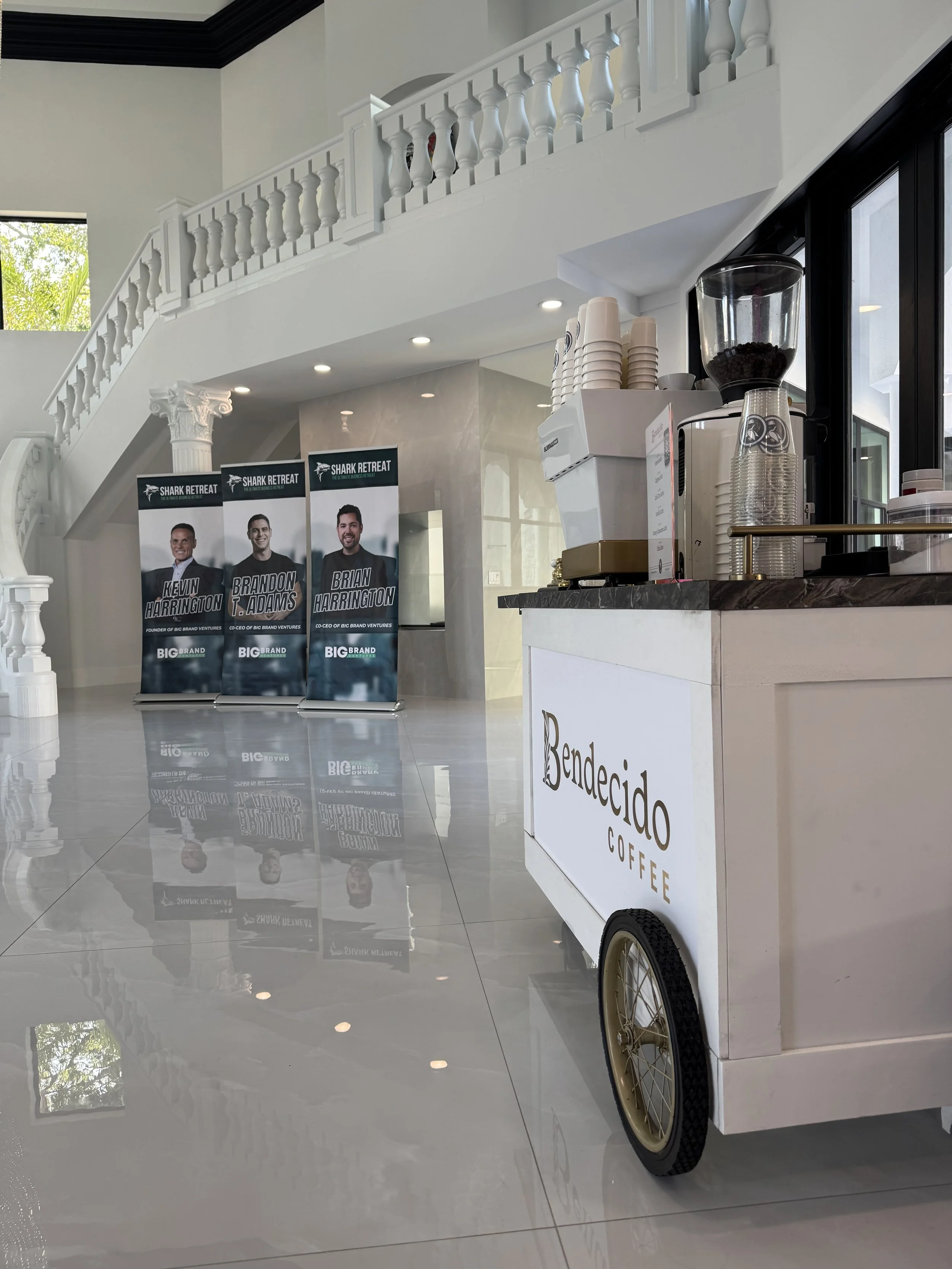 Indoor coffee station with a coffee machine, cups, and a display of promotional banners featuring three men at the Shark Retreat event in South Tampa. White tiled floor and elegant white interior architecture.