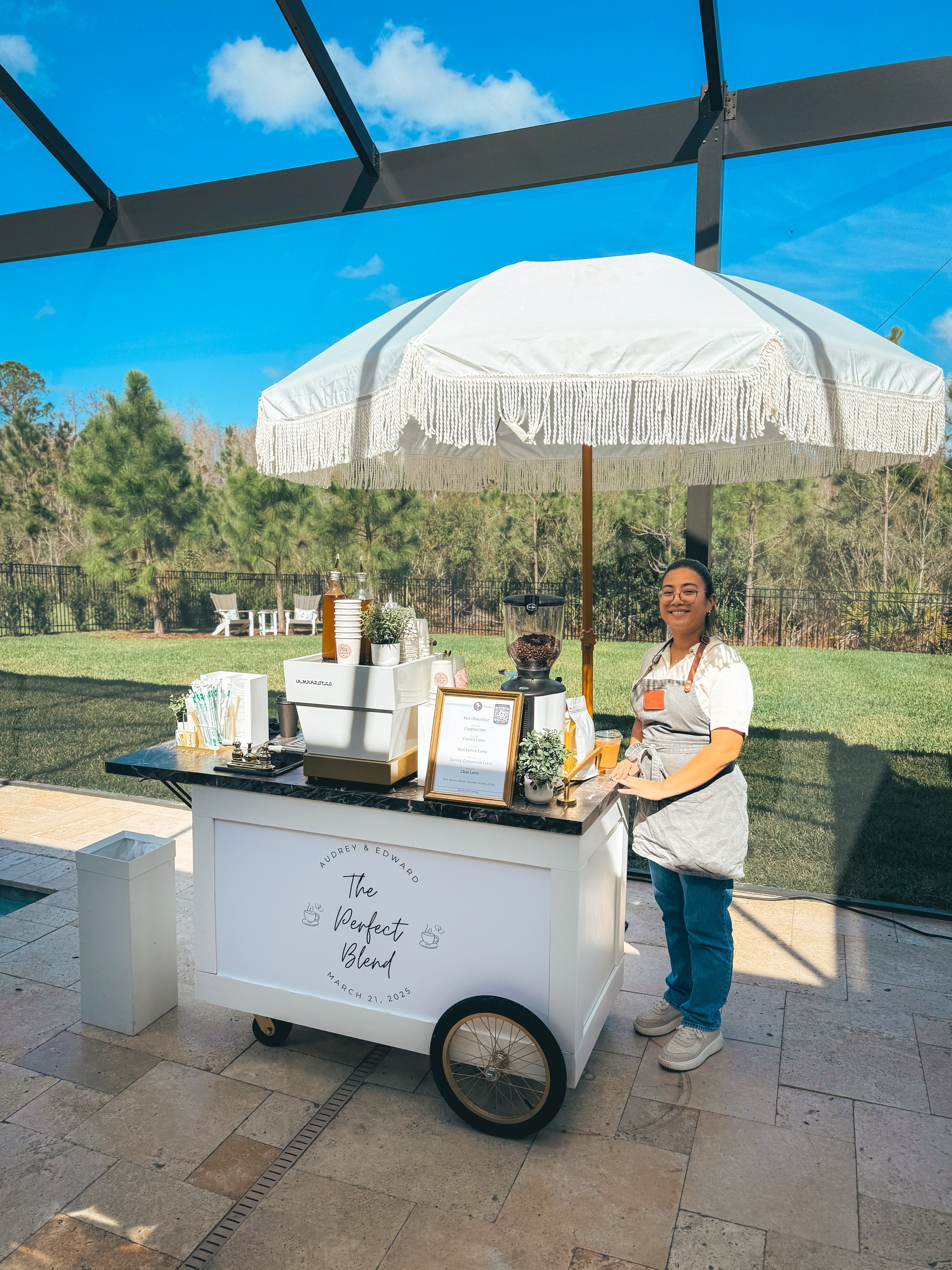 A woman stands behind a coffee cart labeled 'The Perfect Blend' with the date March 21, 2025, on a patio under a large white umbrella. The cart displays coffee supplies, a framed menu, and a coffee grinder. In the background, there is a grassy area, 