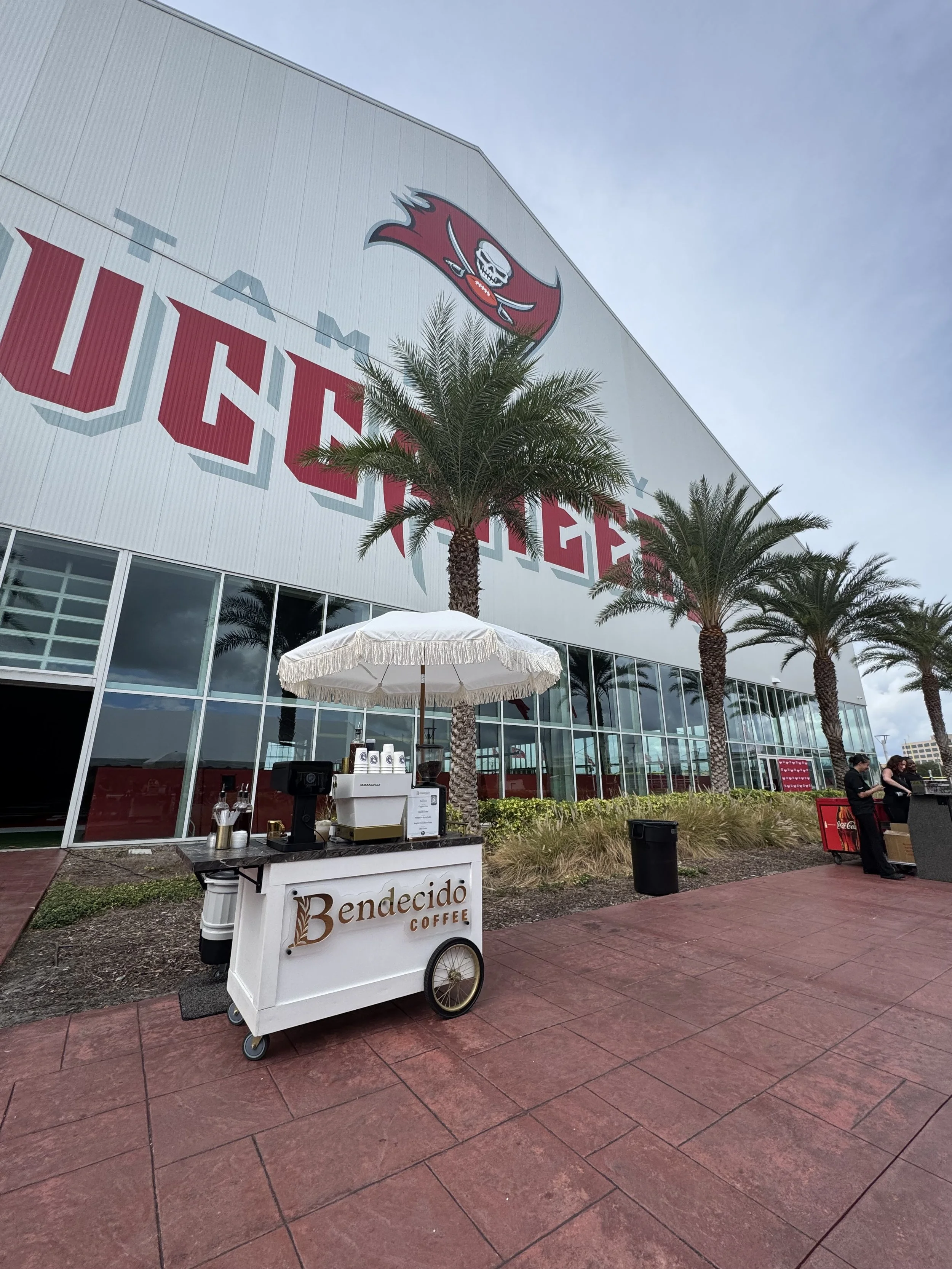 Outdoor coffee stand with umbrellas situated outside a large Tampa Bay Buccaneers arena. Tall palm trees are in front of the arena, which has the team’s logo and name on its facade.