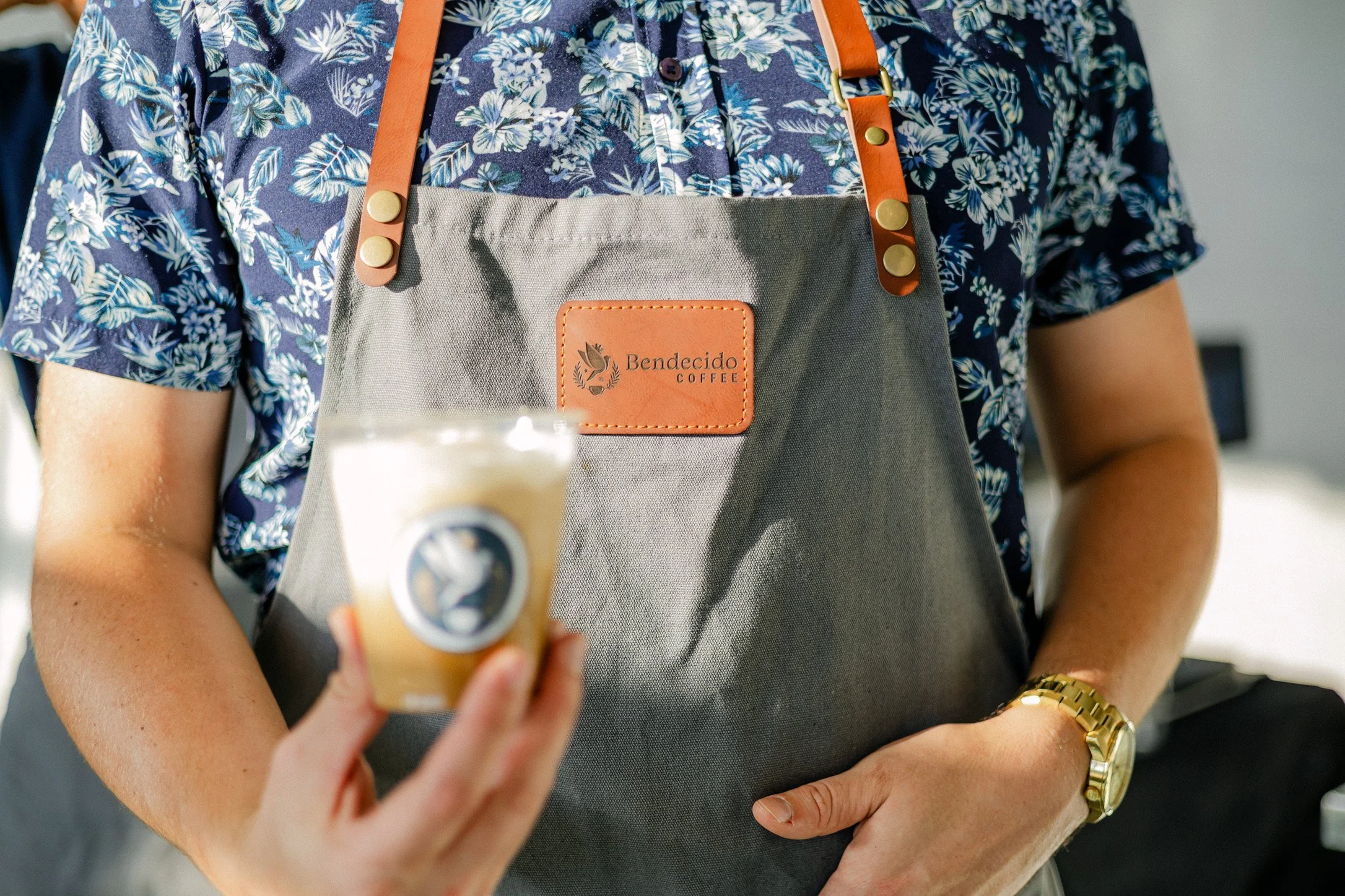 Bendecido Coffee team member in a branded apron holding an iced coffee drink