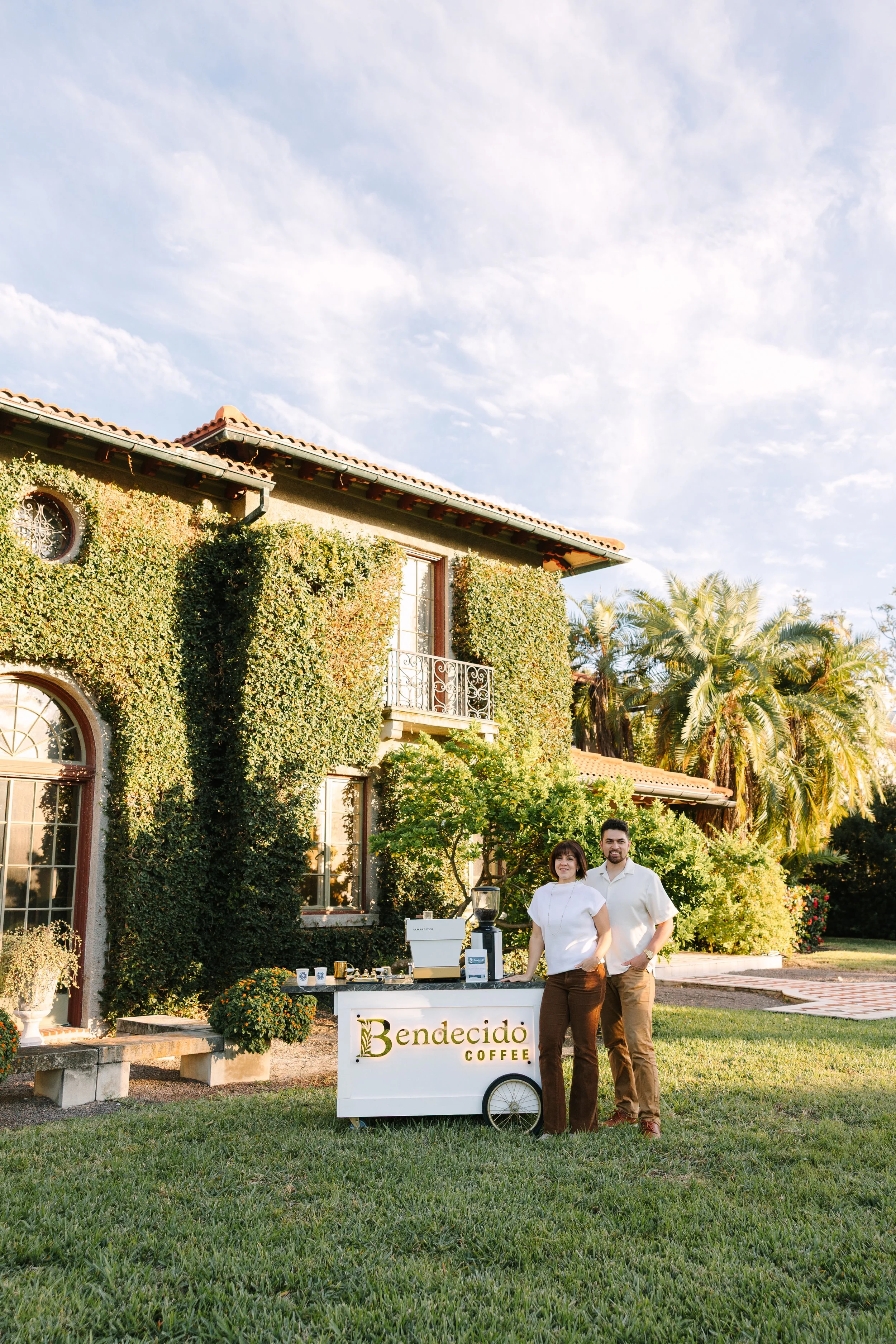 Two people standing next to a coffee cart labeled 'Bendecido Coffee' outdoors in front of a large house covered in ivy, with lush greenery and palm trees in the background during the daytime.