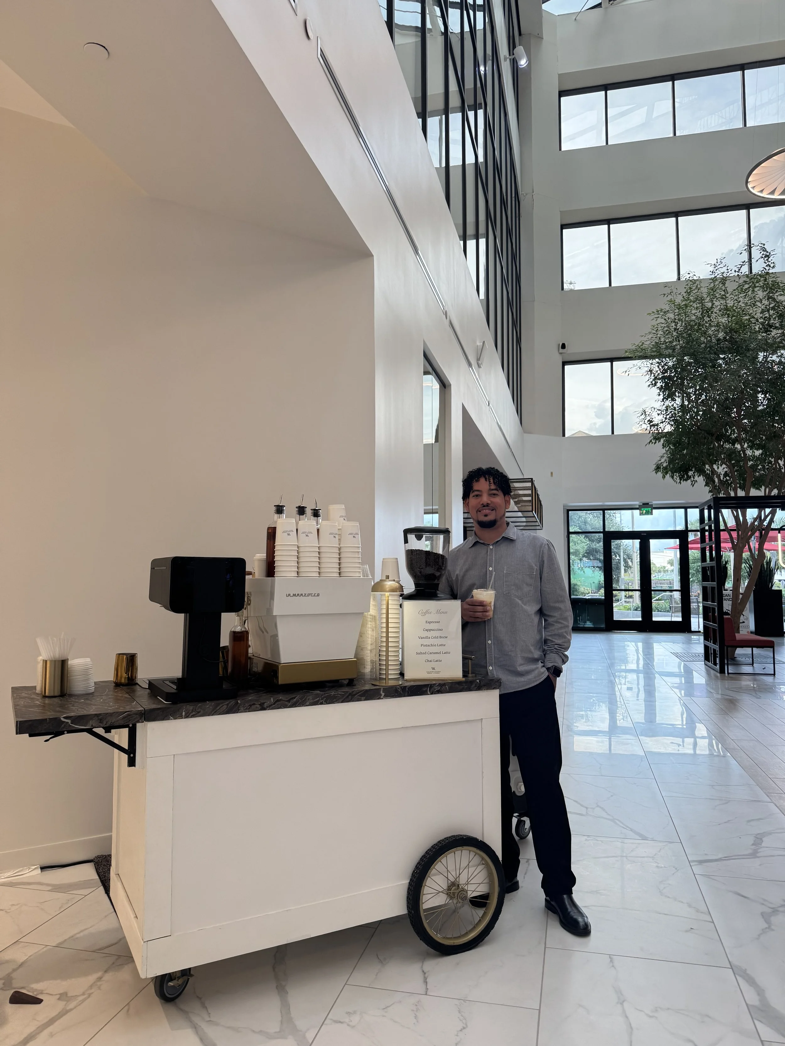 A man standing next to a coffee cart in a modern, spacious lobby, holding a drink and smiling. The coffee cart has a coffee machine, cups, and a menu sign. Large windows and high ceilings are visible in the background.