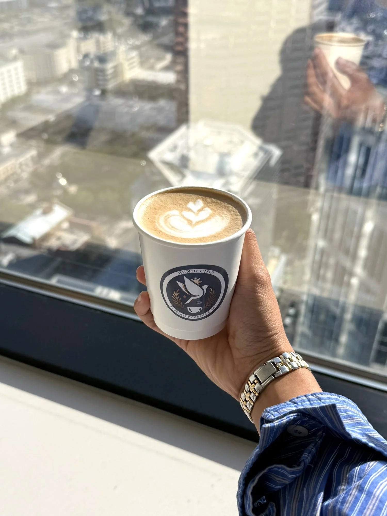 Latte with Bendecido Coffee logo cup held up to a high-rise window above a downtown Tampa street