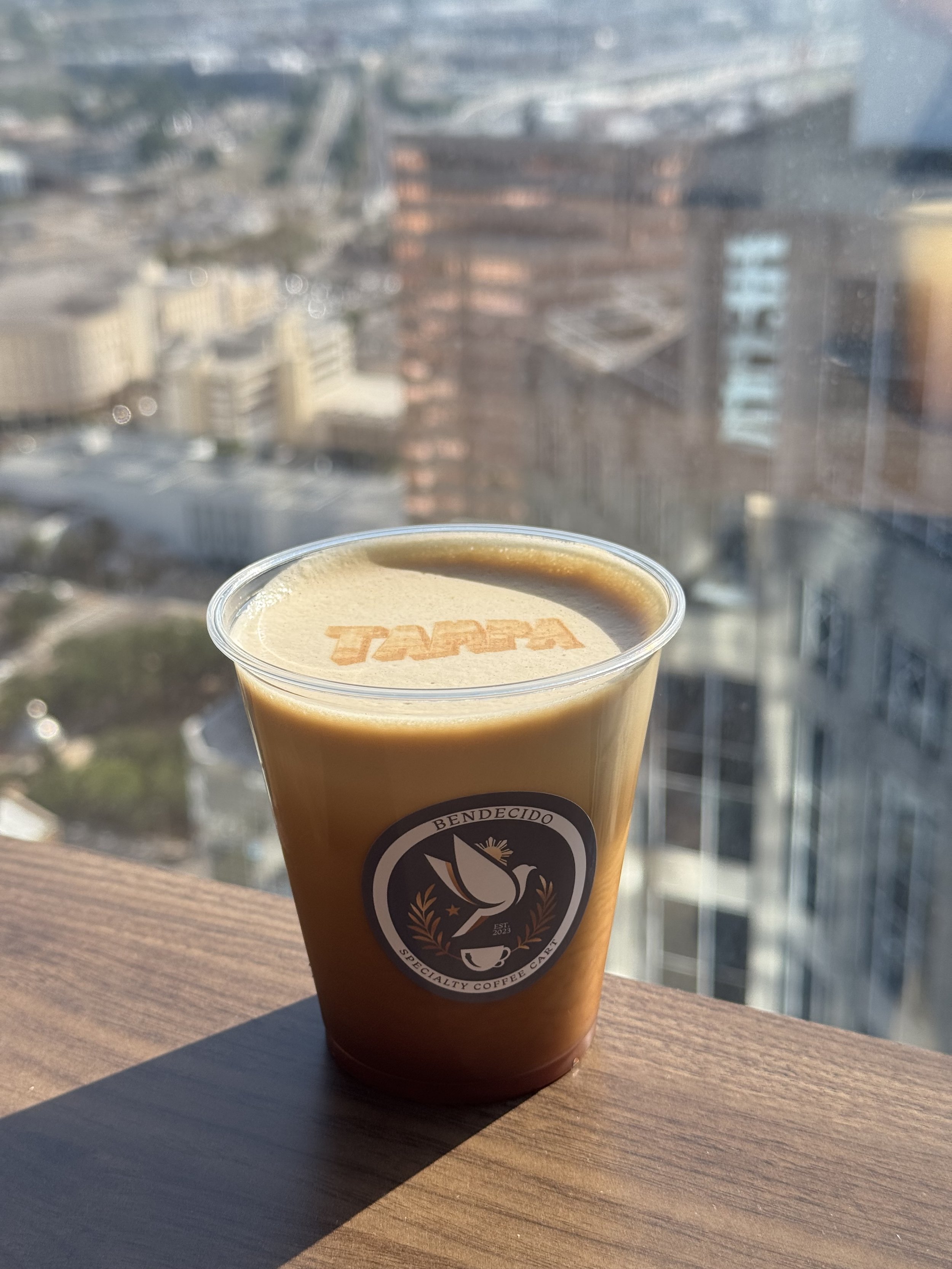 A coffee cup with a logo that says 'Bendecido Specialty Coffee Cafe' on a wooden surface with a cityscape view in the background.