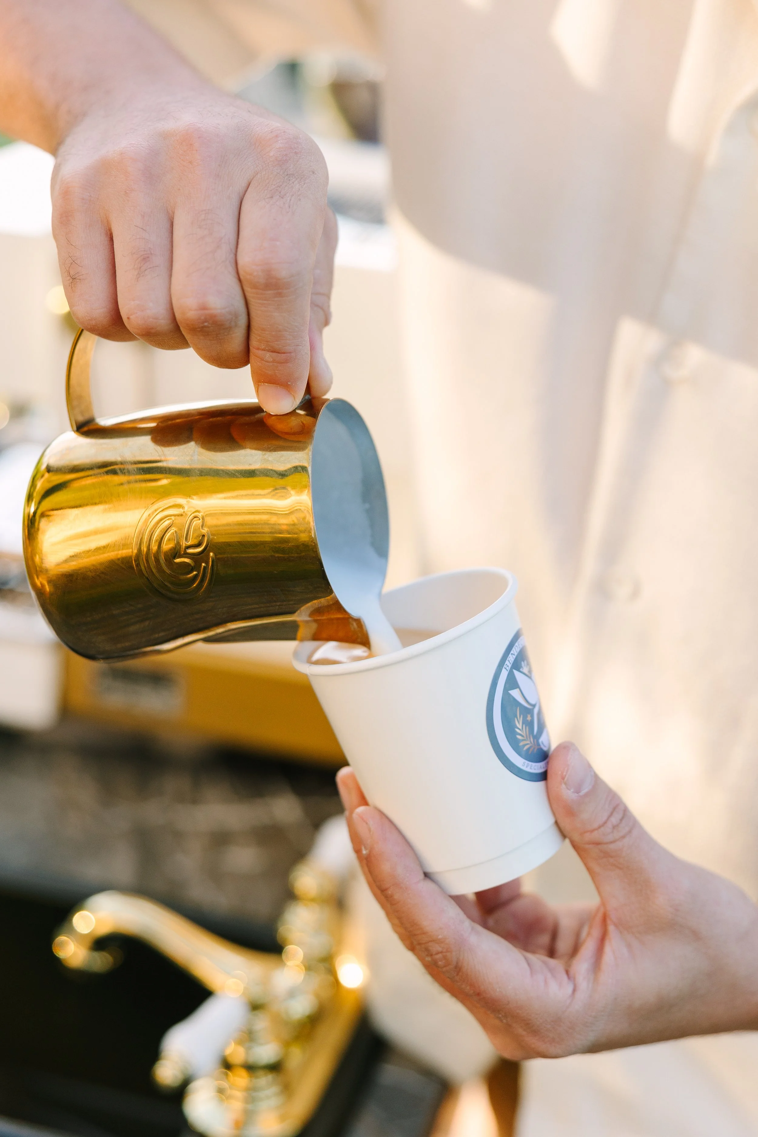A person pours milk froth from a golden metallic pitcher into a paper cup with a logo on it.