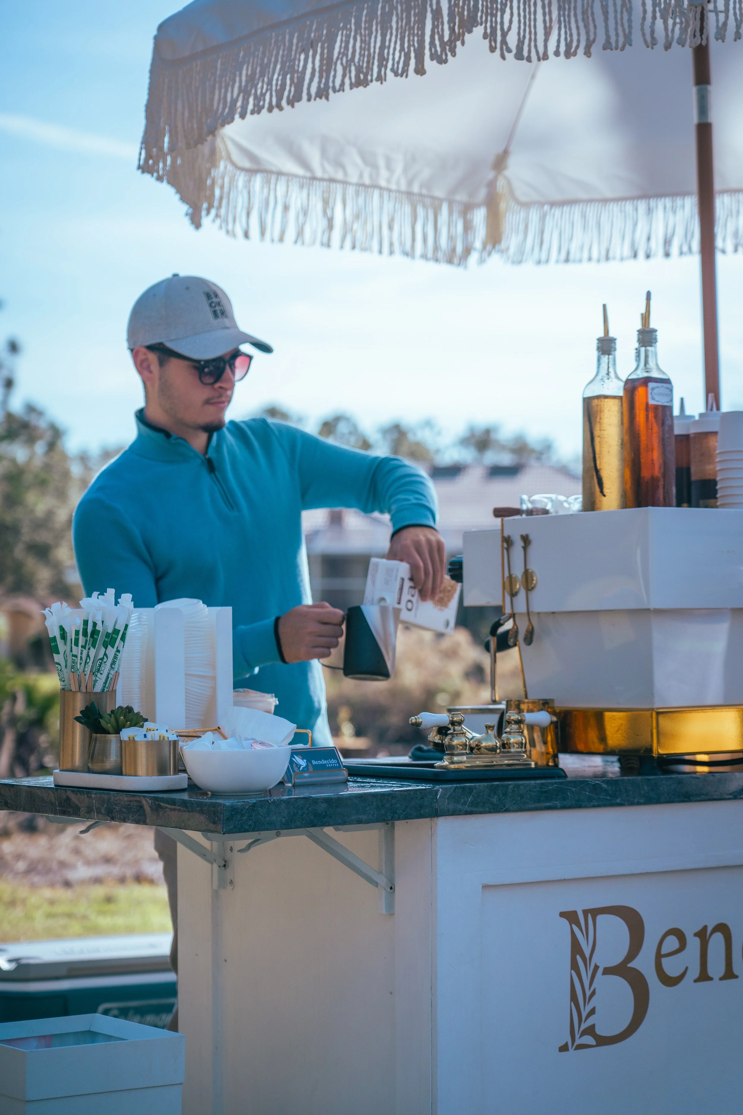 A man at a beverage stand under a large umbrella, pouring a drink from a carton into a cup. The stand has bottles of syrup and disposable cups.