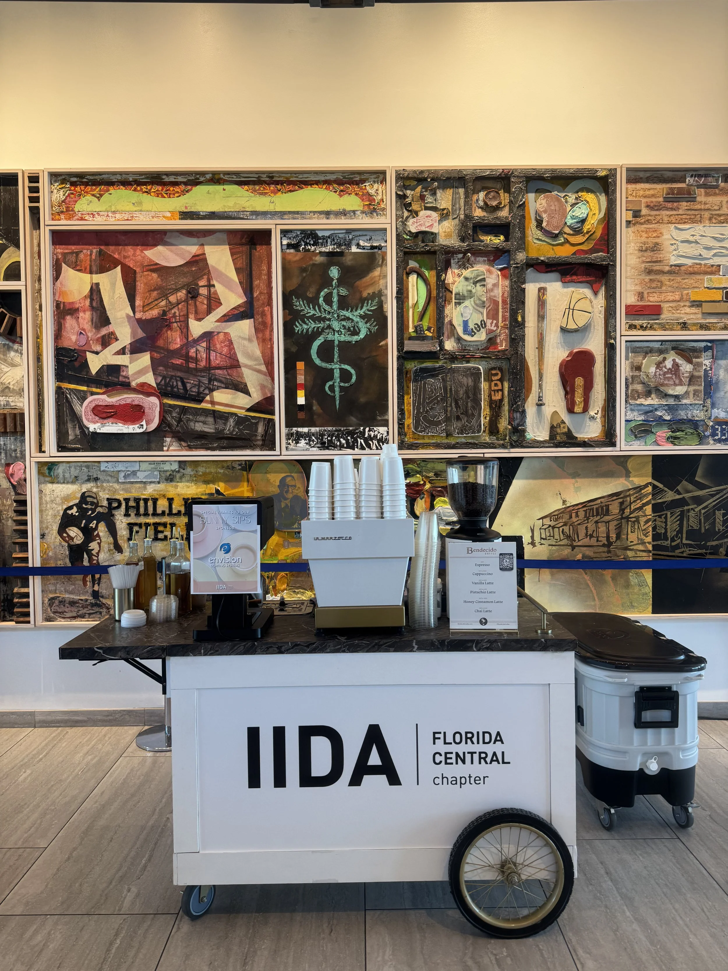 Coffee stand with IIDA Florida Central Chapter sign, featuring cups, a coffee grinder, and menu, set against a colorful mural at the Tampa River Center.