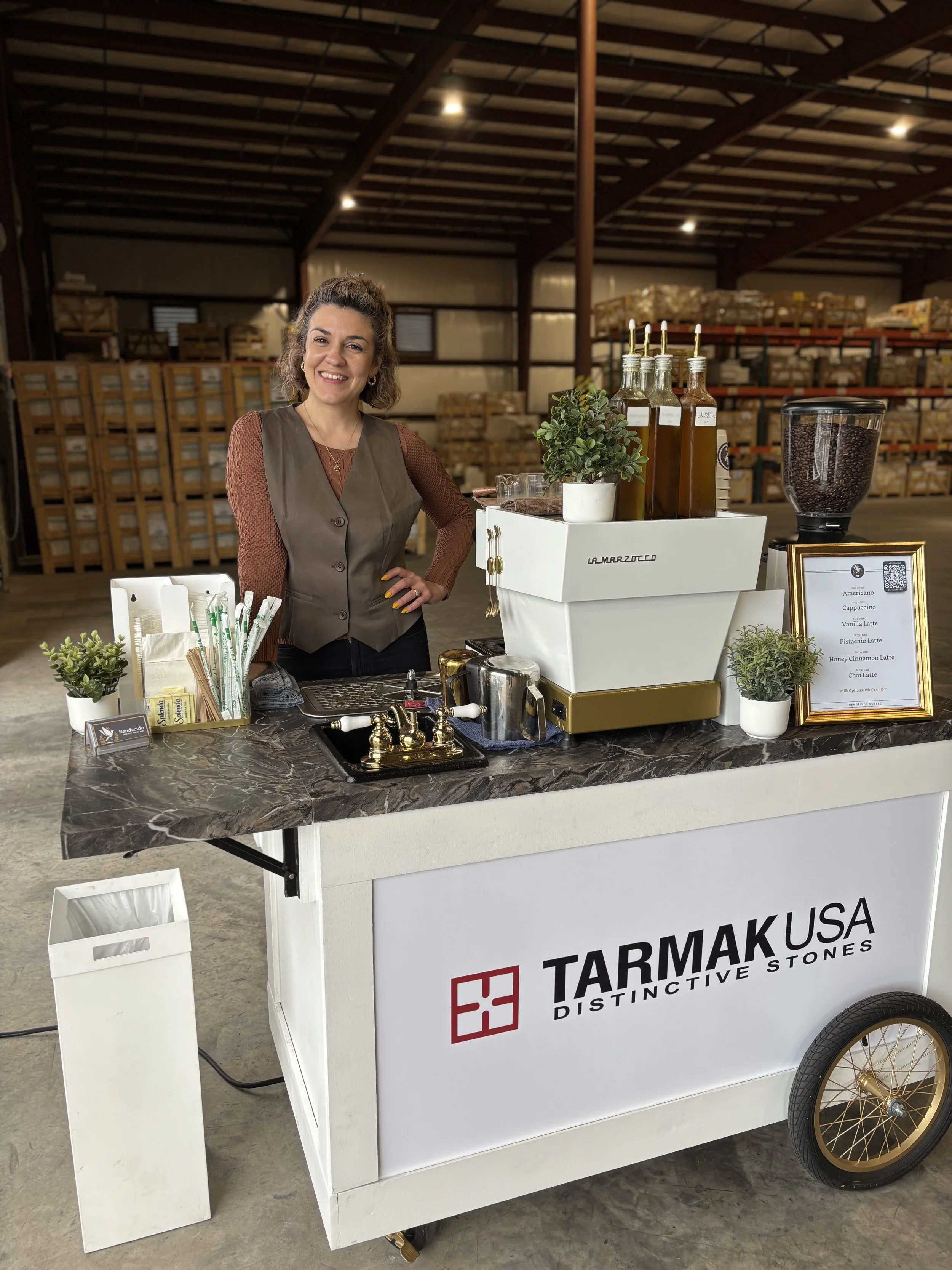 Smiling woman standing behind a portable coffee cart with bottles, potted plants, a framed menu, and coffee beans in a Tampa warehouse setting.