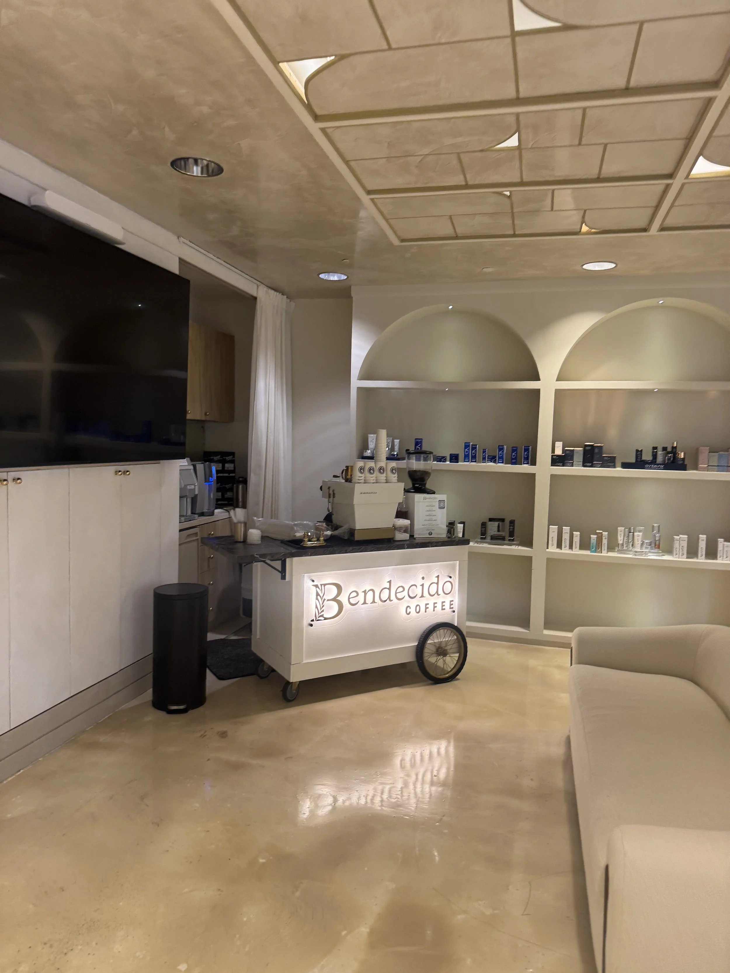 Inside a coffee shop with a coffee cart labeled 'Bendecido Coffee,' shelves with coffee products, a large television, and a beige sofa.