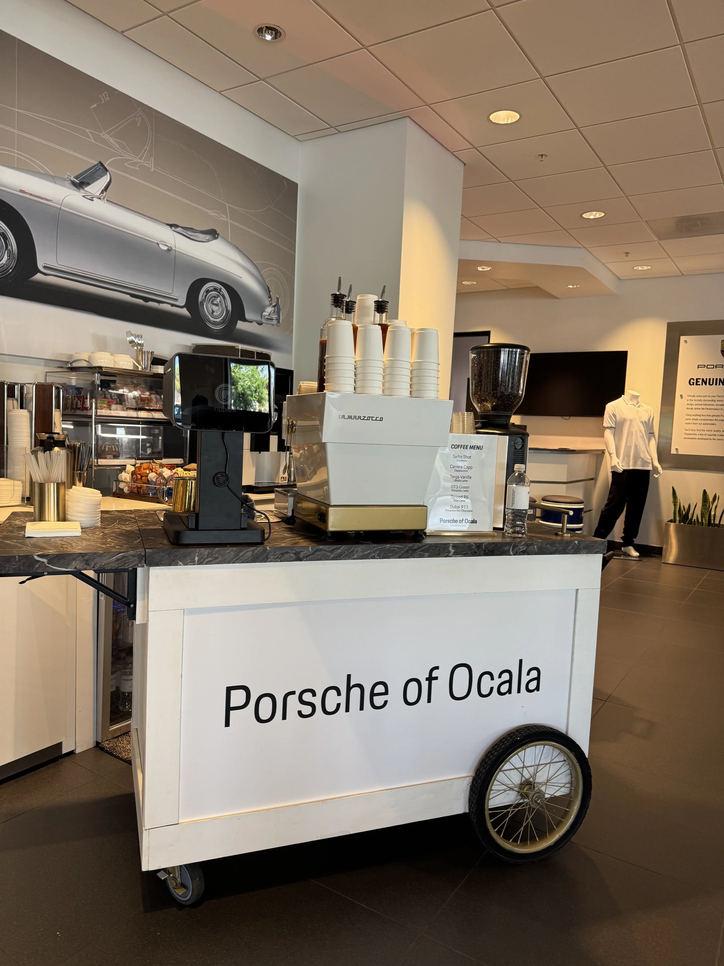Coffee cart with 'Porsche of Ocala' sign, positioned inside a showroom with a large car poster on the wall and a mannequin in the background.
