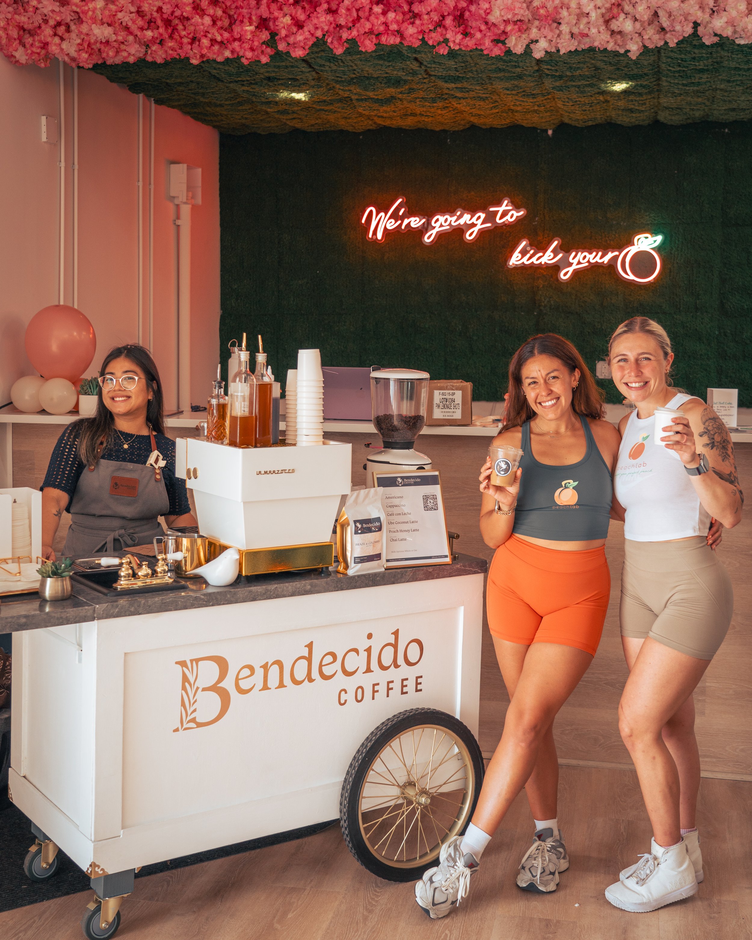 Three smiling women at a coffee cart labeled 'Bendecido Coffee', with a neon sign reading 'We're going to kick your 🍑' in the background and flowers on the ceiling.