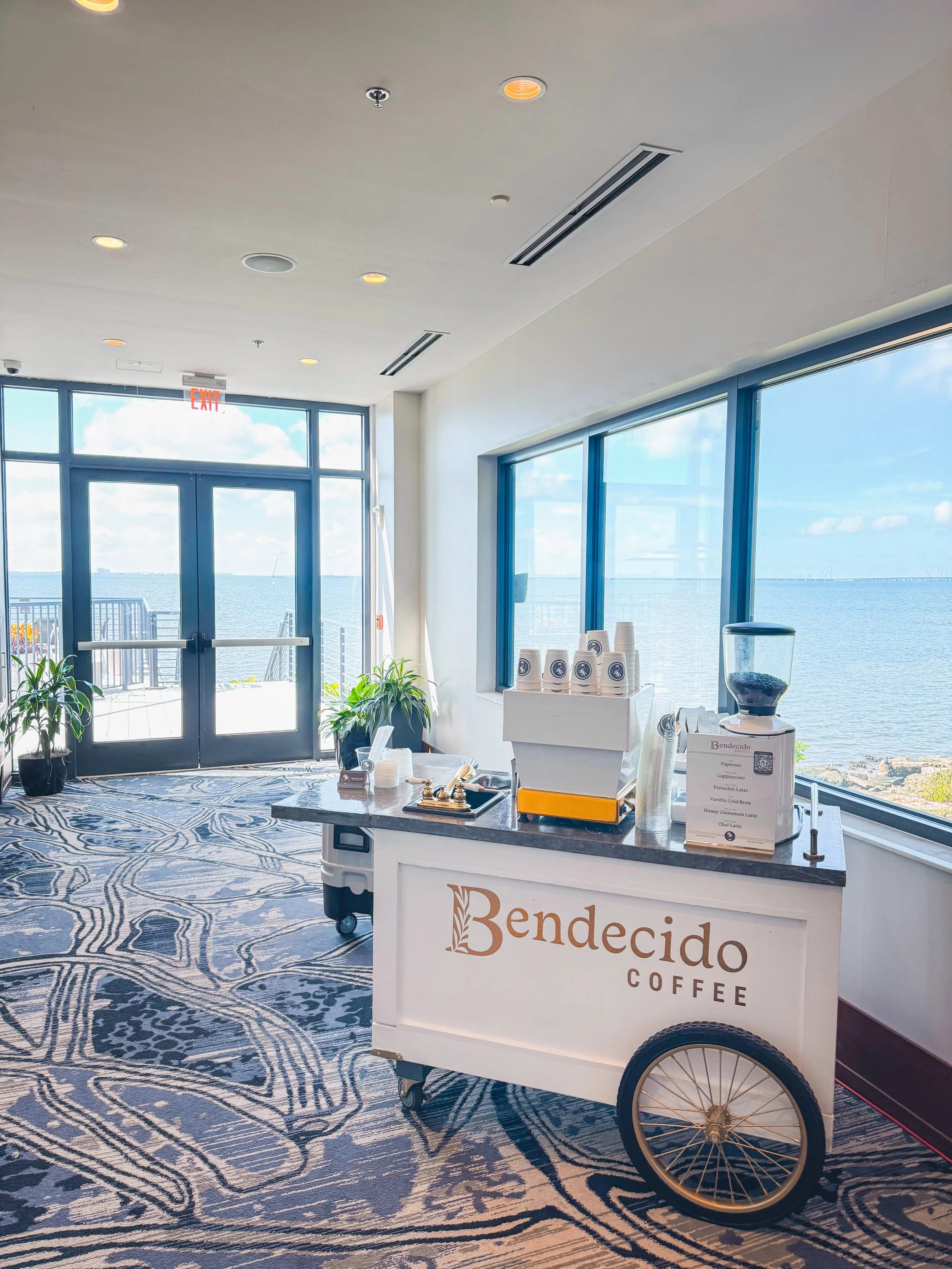 Coffee cart with 'Bendecido Coffee' logo, set near large windows overlooking water, with doors and potted plants in the background.