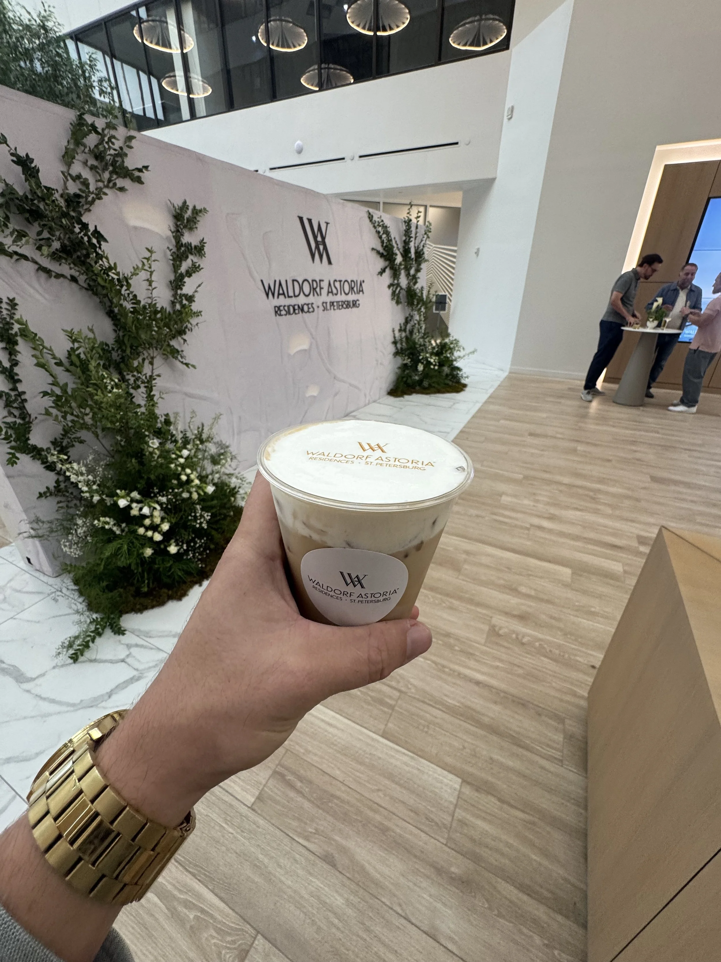 Person holding a coffee drink with a Waldorf Astoria logo in a modern lobby with a decorative wall and people in the background.