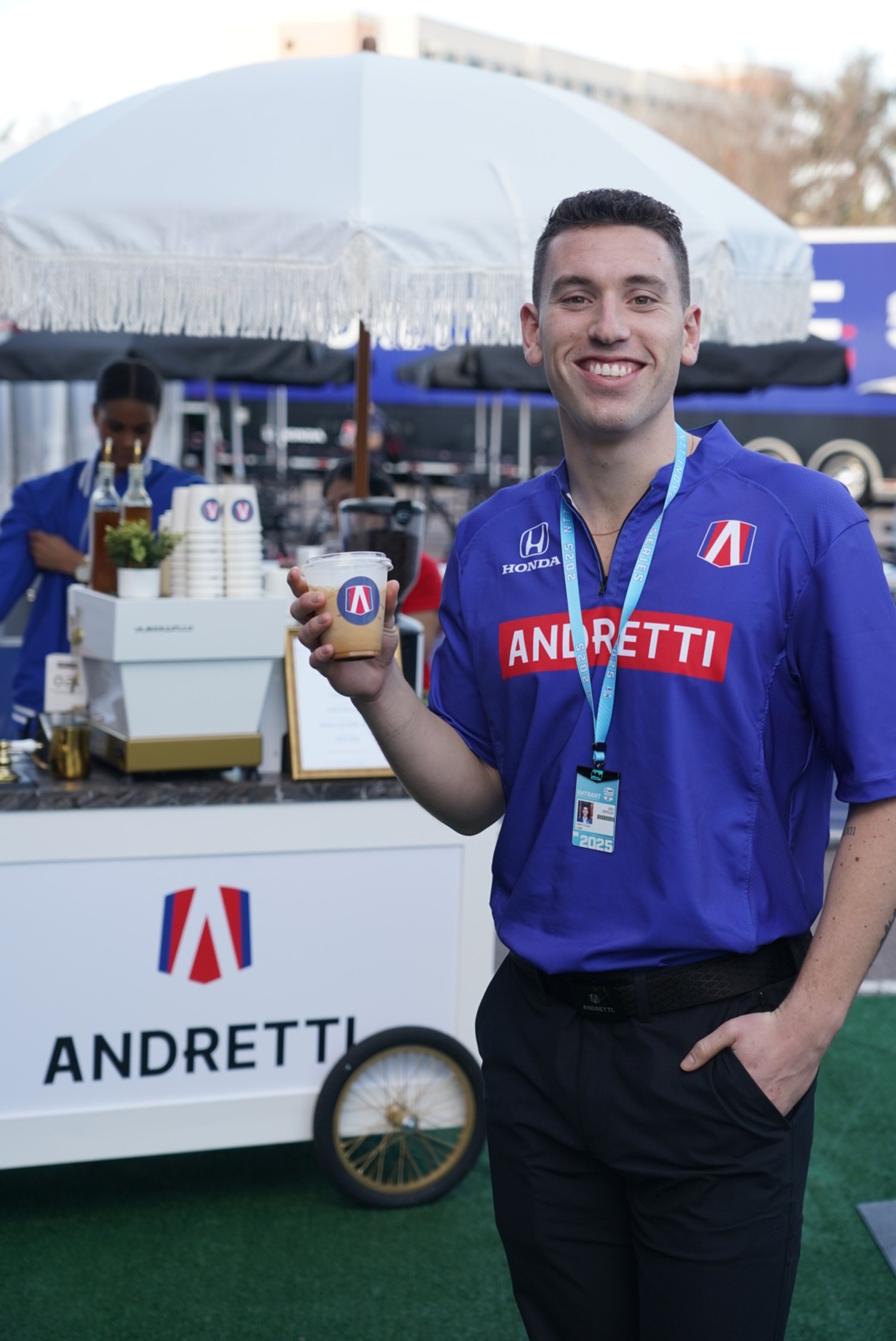 A man in a blue racing team shirt smiling and holding a beverage with an Andrettis logo, standing in front of a beverage stand with an umbrella, a woman serving drinks in the background at the St. Pete Grand Prix.
