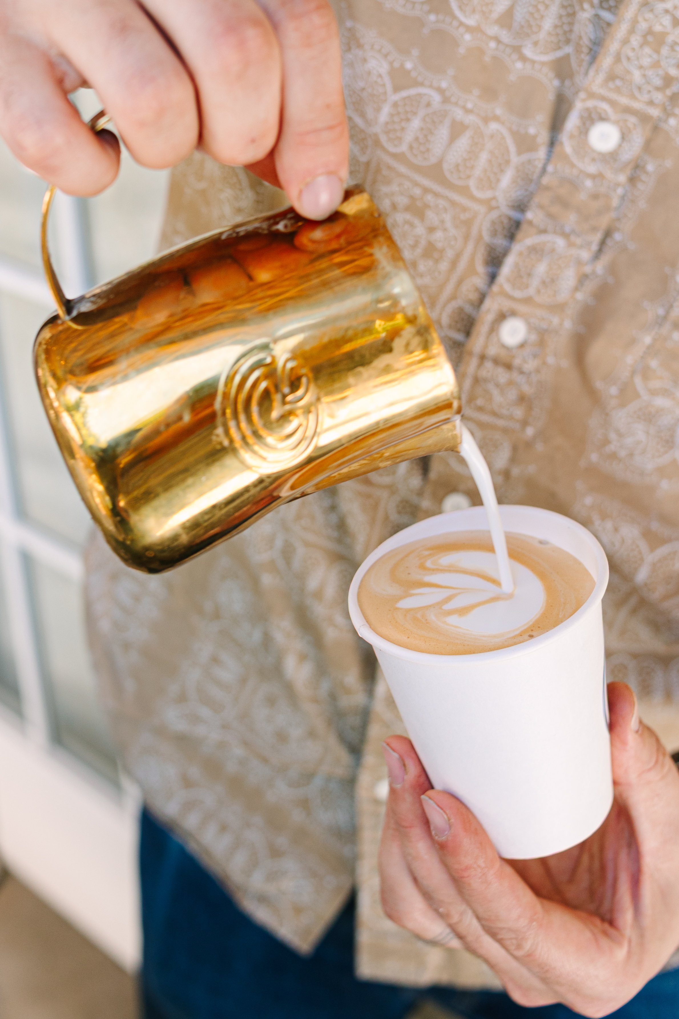 A Bendecido Coffee barista pours coffee from a shiny gold mug into a white disposable coffee cup with latte art on top. The person is wearing a patterned shirt and a ring on their hand.