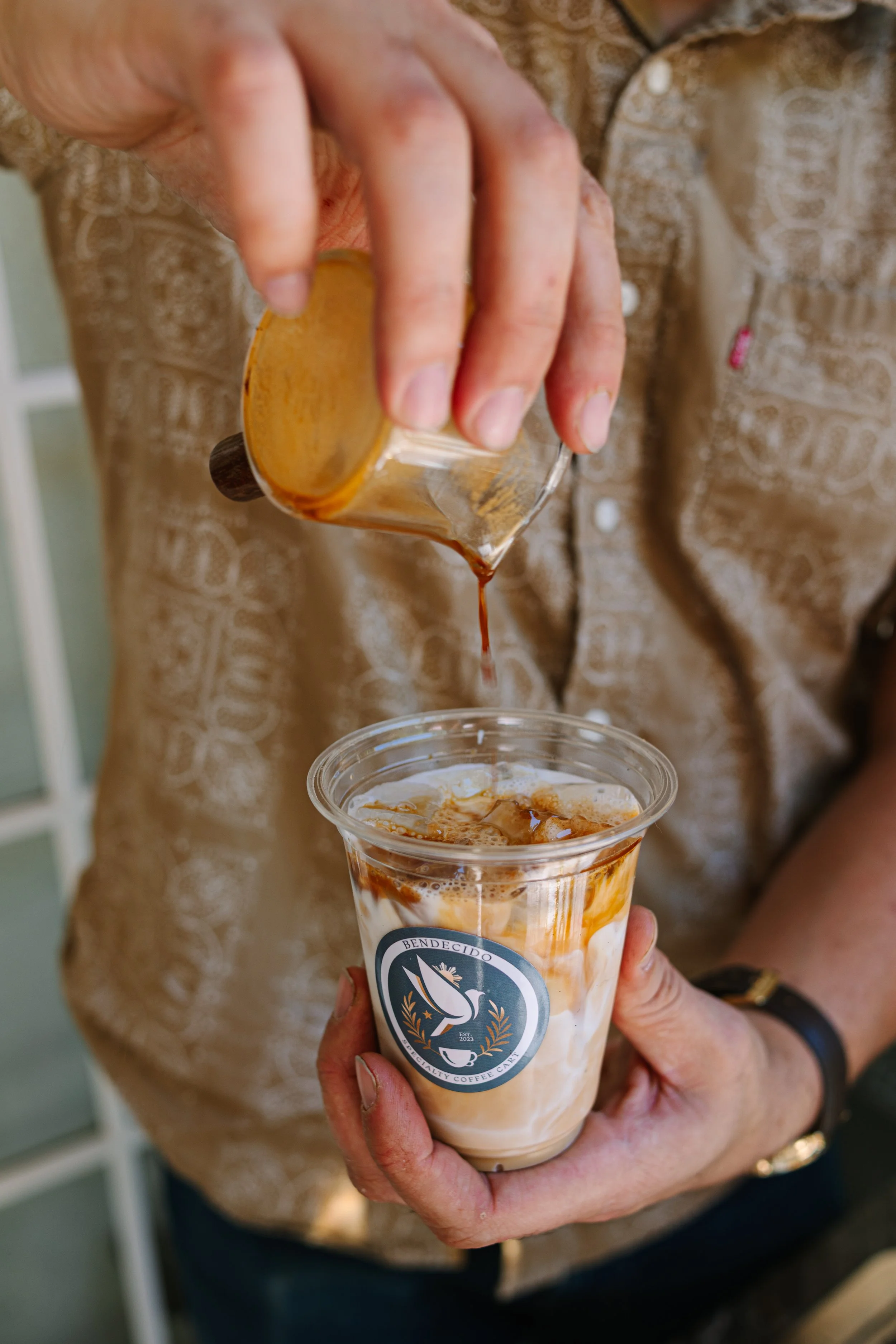 A person pours a caramel syrup into an iced Bendecido coffee drink with cream in a plastic cup, held with one hand, with a logo visible on the cup.