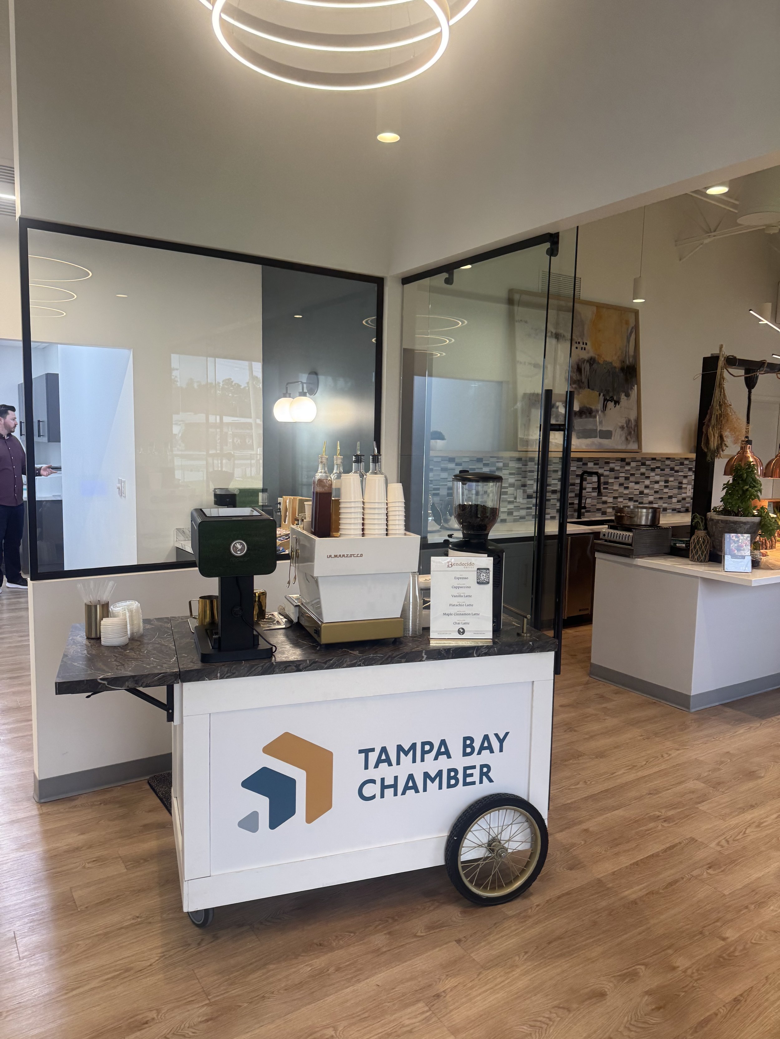 Coffee cart with Tampa Bay Chamber logo, coffee cups, syrup bottles, coffee grinder, and menu sign, set up inside a modern, well-lit café with wooden floors and a glass partition.