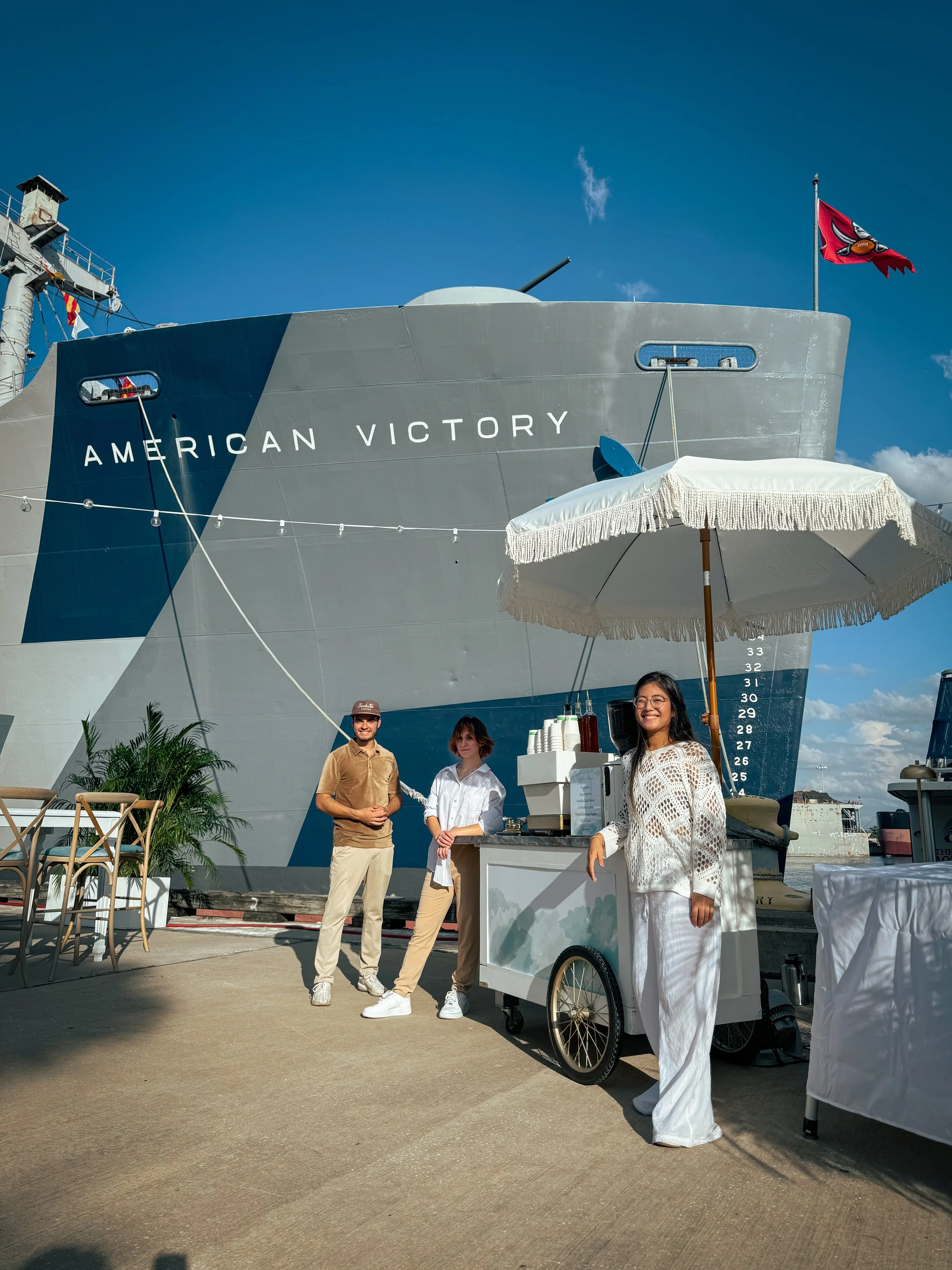 People standing in front of a large ship named American Victory at a dock, with a small ice or drink cart and an umbrella in the scene.