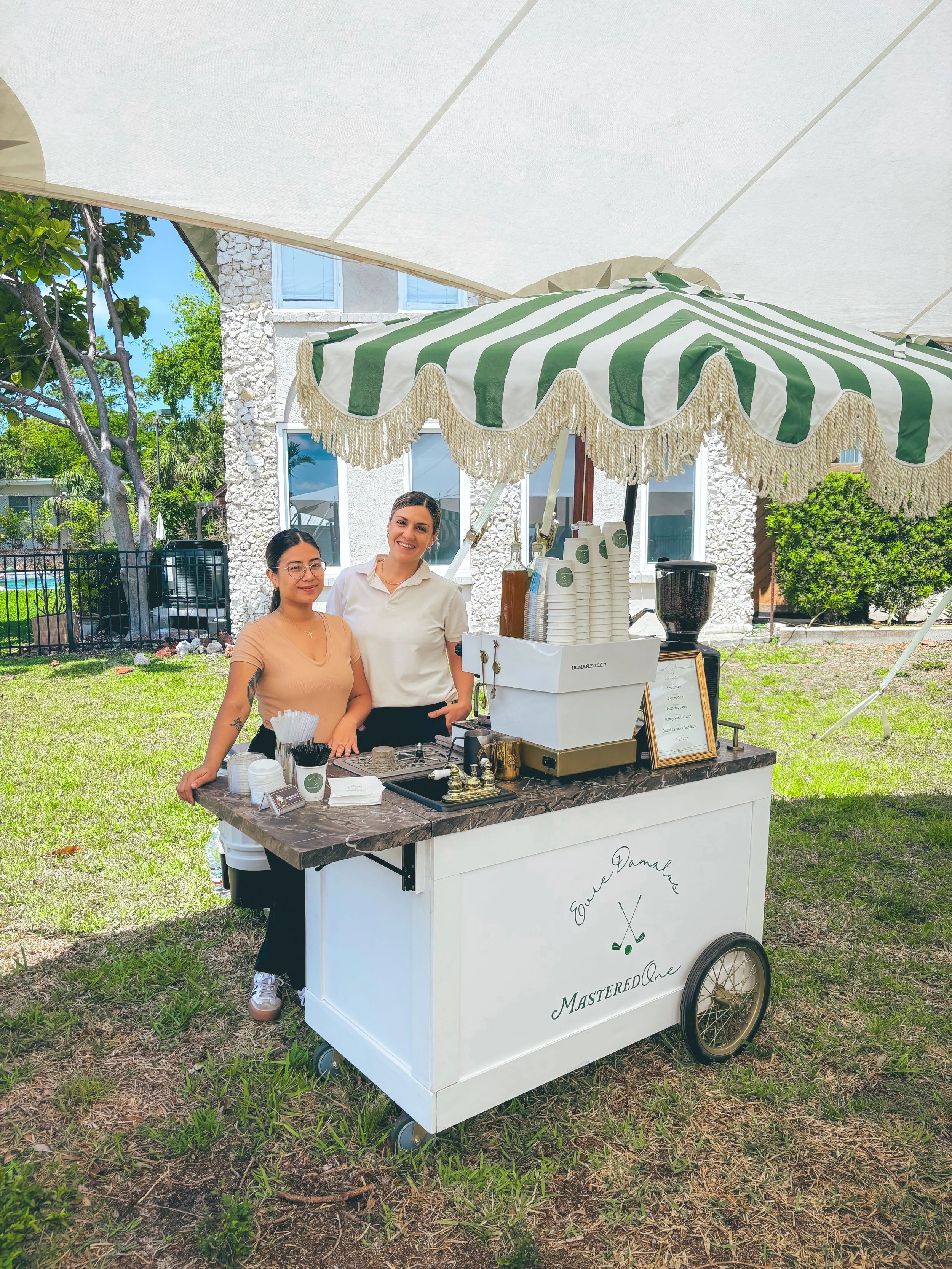 Two women standing behind a mobile ice cream or coffee cart under a large umbrella in a backyard with grass and trees.