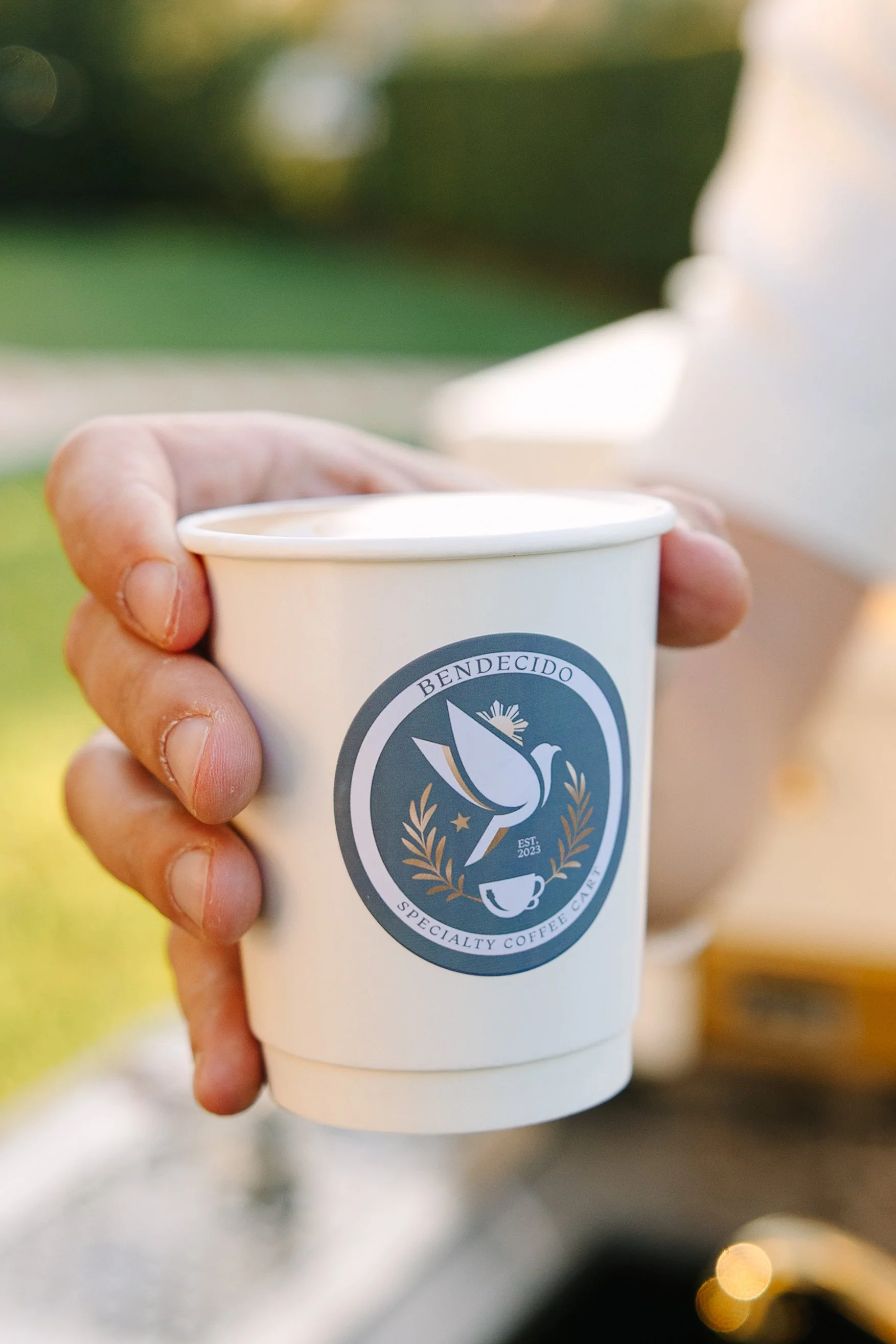A hand holding a white coffee cup with a logo featuring a dove, laurel wreath, star, and coffee cup, labeled "Bendecido Specialty Coffee Cafe."