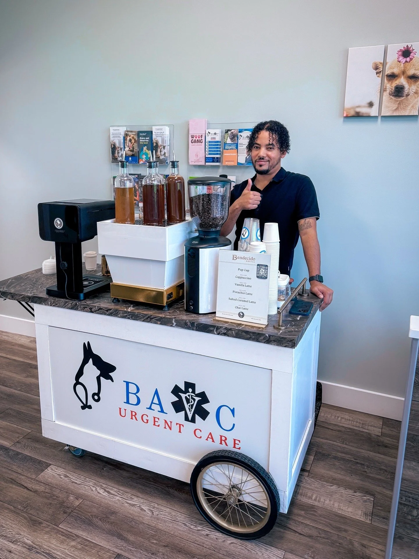 A man standing behind a mobile coffee cart labeled 'BADC Urgent Care', serving coffee drinks on a marble countertop, with bottles of flavored syrups, a coffee grinder, and cups.