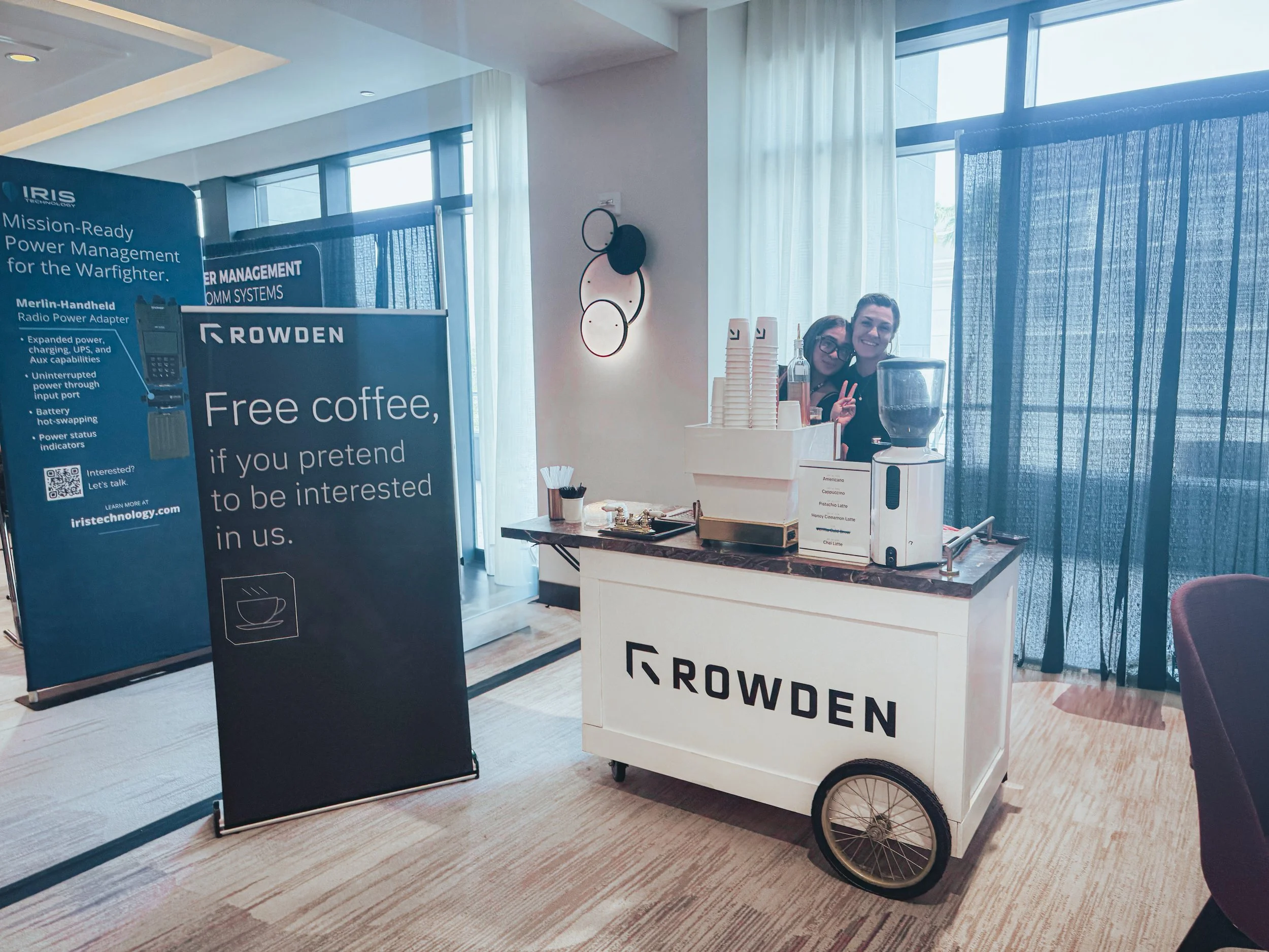 A mobile coffee station with two smiling women behind it, signboard offering free coffee, blue curtains, and natural lighting from windows.