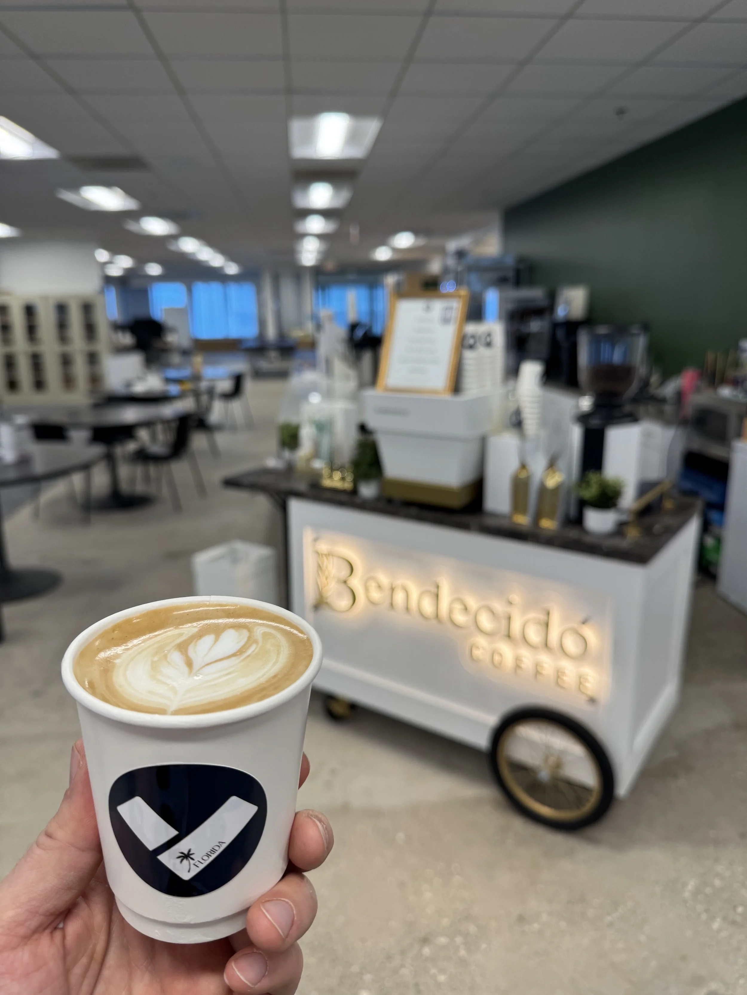 A hand holding a paper cup topped with latte art in front of a coffee cart with a sign that reads 'Bendejado Coffees' in an Downtown St Pete office.