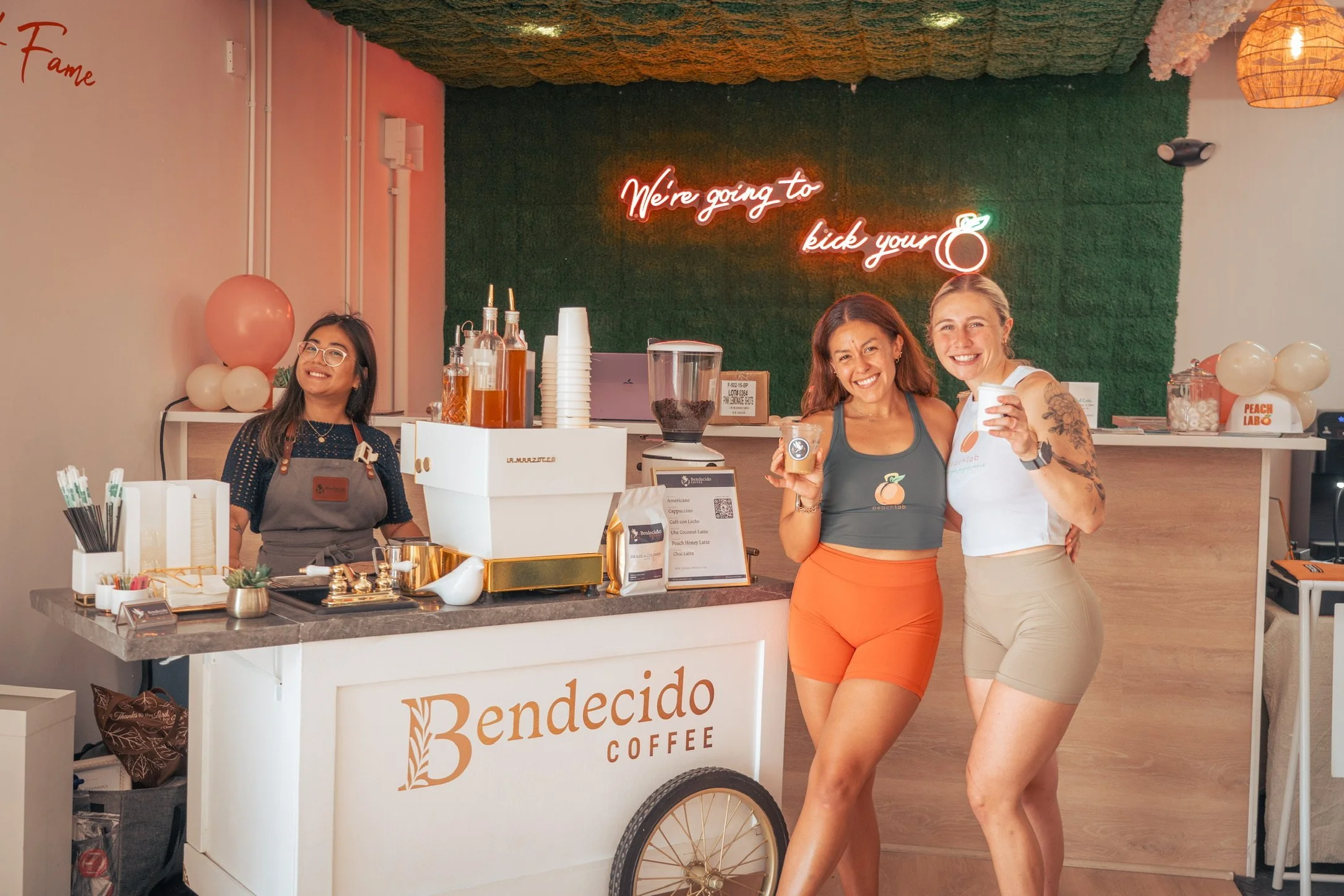 Three smiling women posing behind a Bendecido Coffee cart under a neon sign