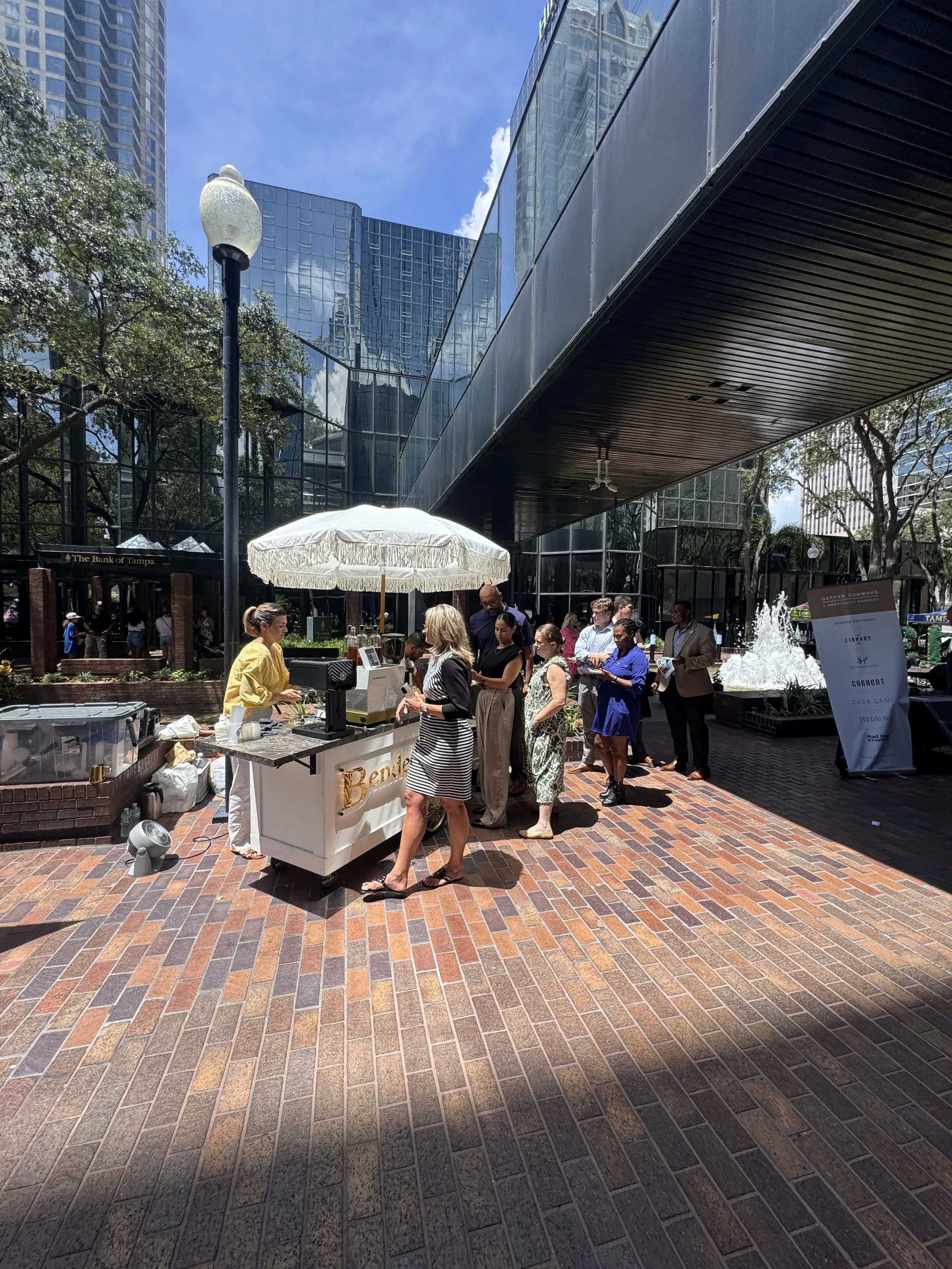 People queueing at a coffee stand outside a modern glass building on a sunny day, with trees, a fountain, and a sign in the background.