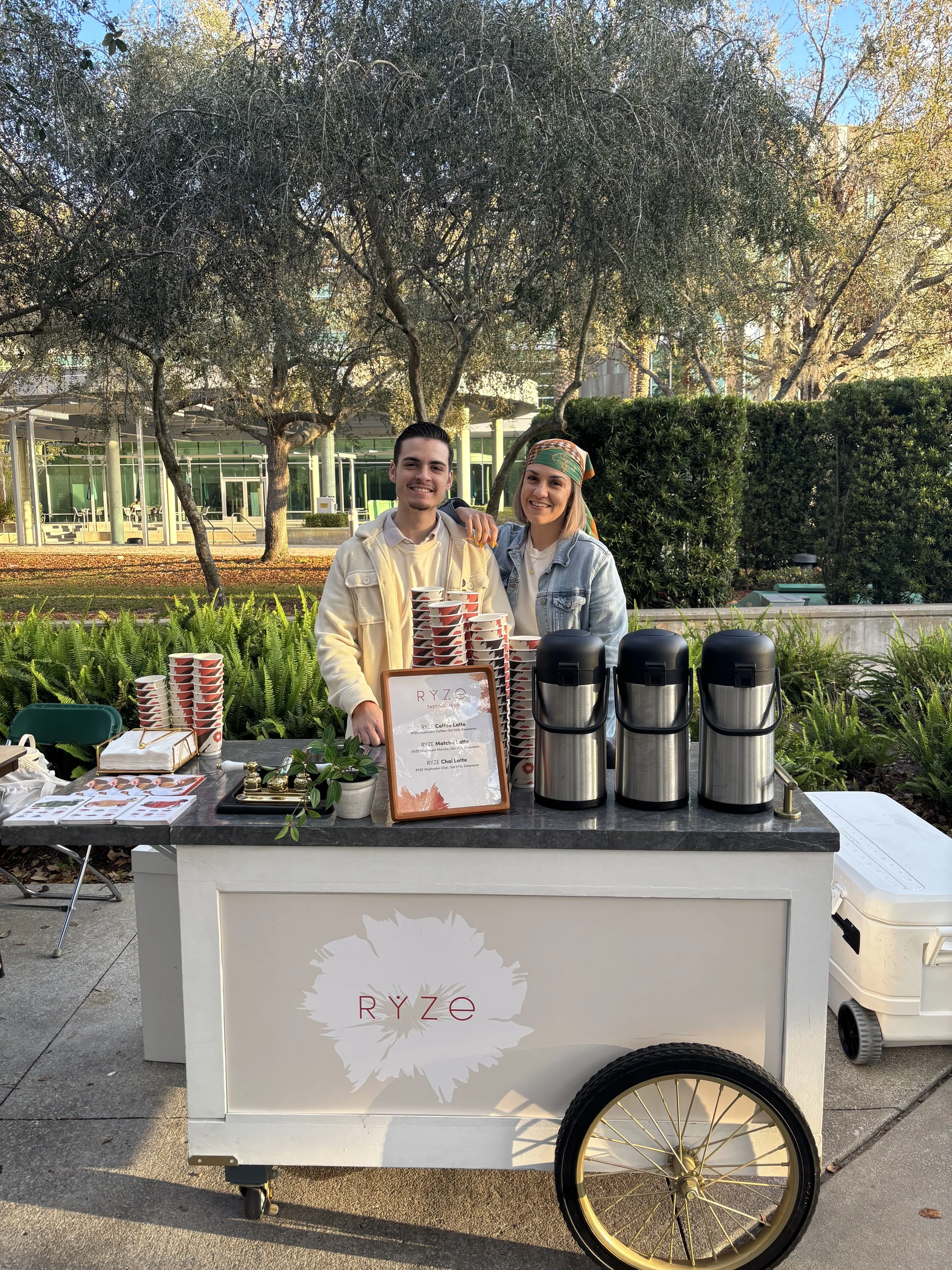 Two people standing behind a coffee cart outdoors, smiling, with trees and a building in the background. The cart has four large coffee dispensers and a menu sign at the University of South Florida.