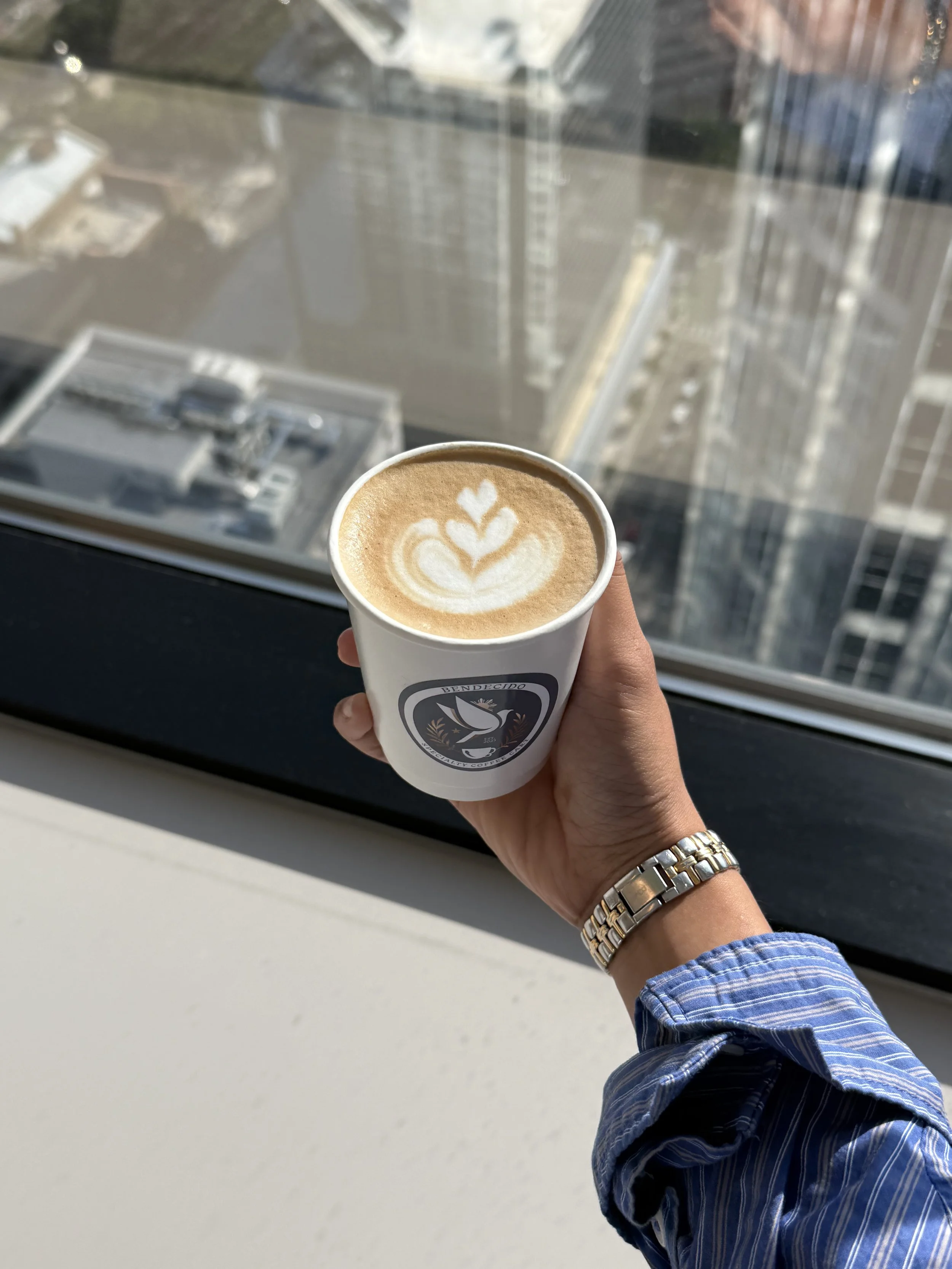 Hand holding a cup of latte art coffee with a city view through a large window in the background.