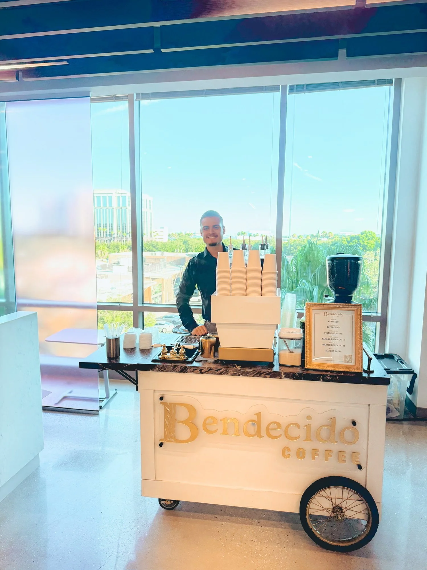 Barista at the Bendecido Coffee cart beside floor-to-ceiling windows at a Tampa Bay office event
