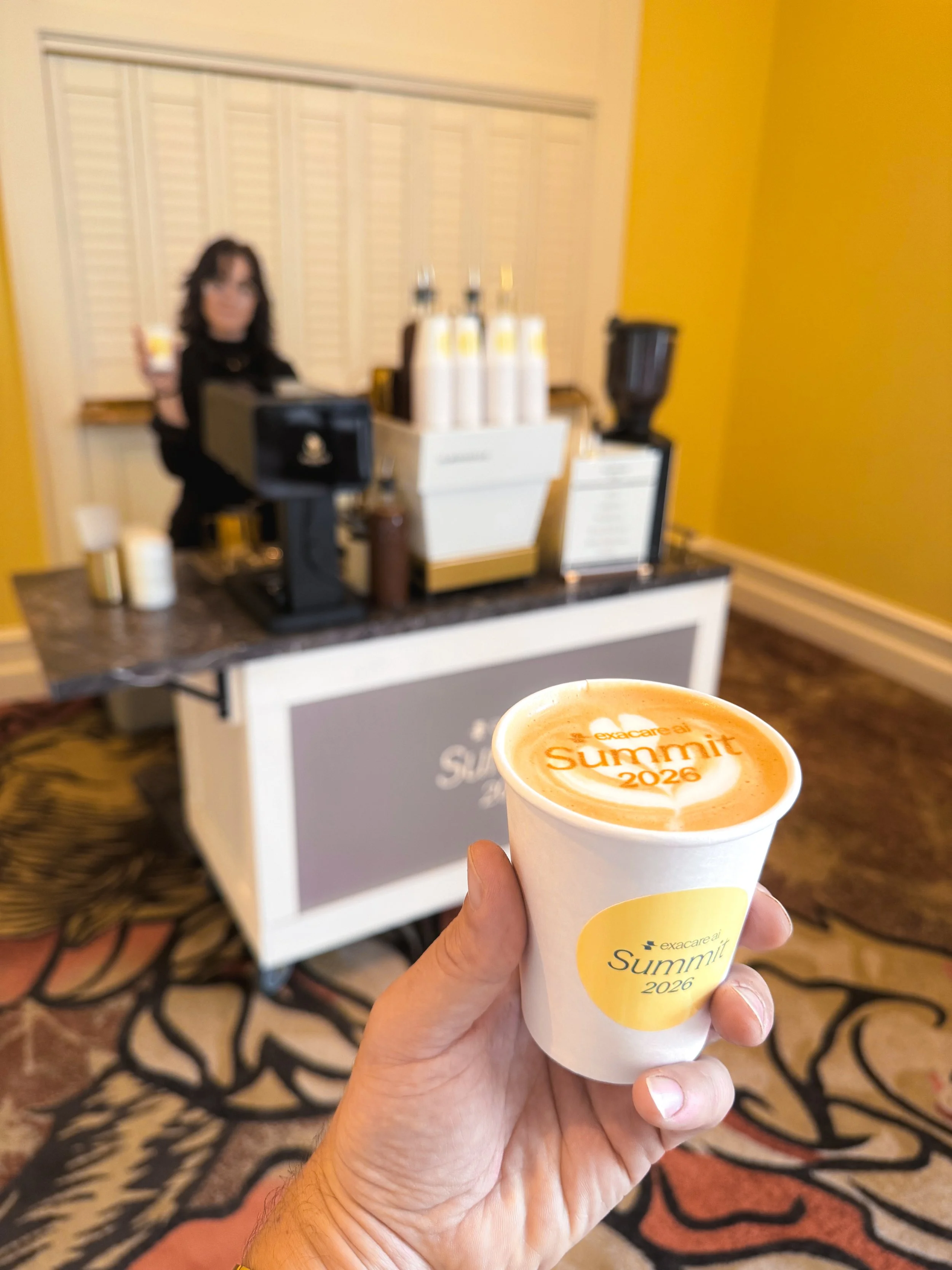 Person holding a cup of Bendecido Coffee with 'excare ai Summit 2026' written on it, inside a conference or hotel lobby with a coffee station in the background.