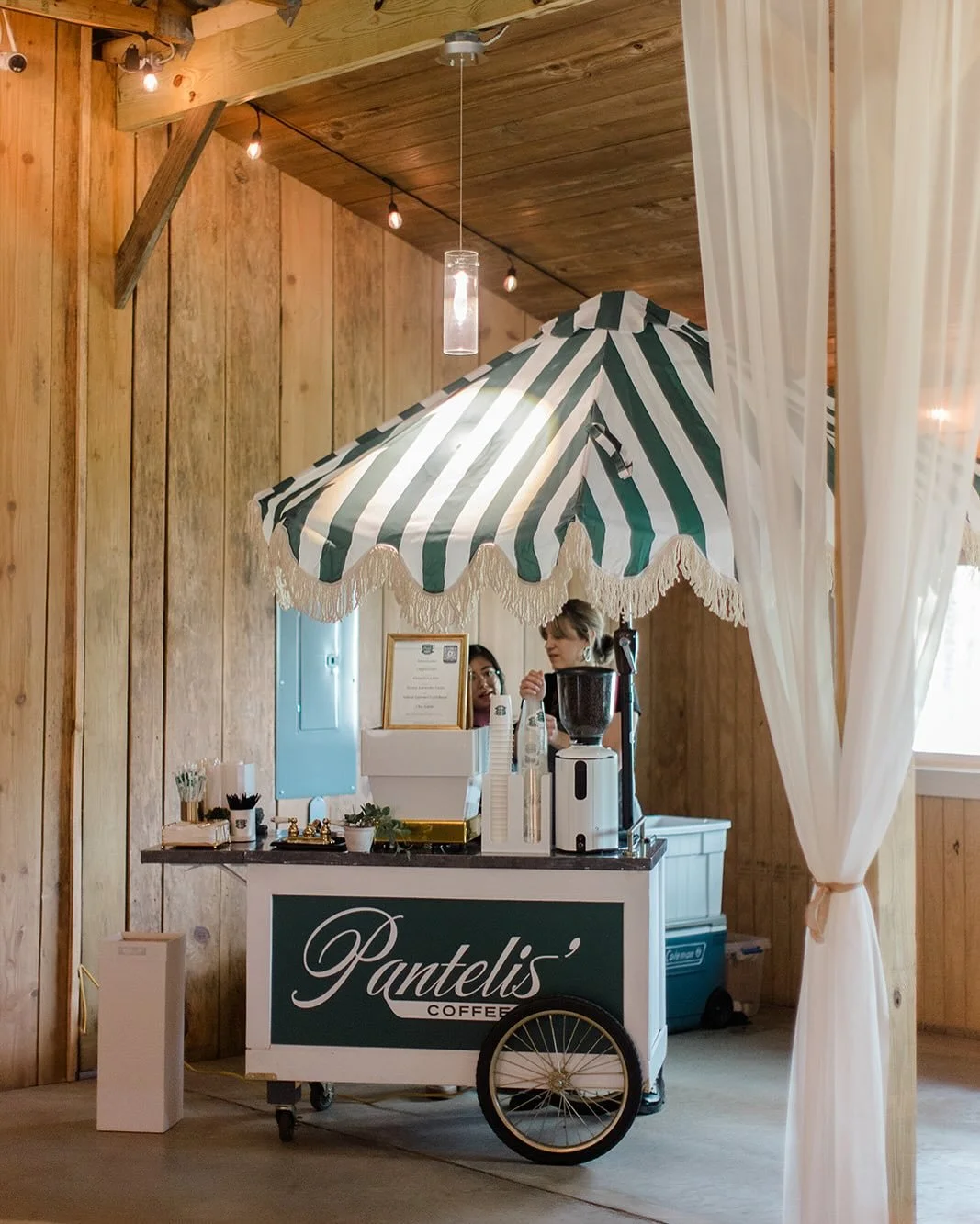 A coffee cart with a green and white striped umbrella, set up indoors against wooden walls. Two people are behind the cart, one serving coffee, with various coffee-making supplies on the cart.