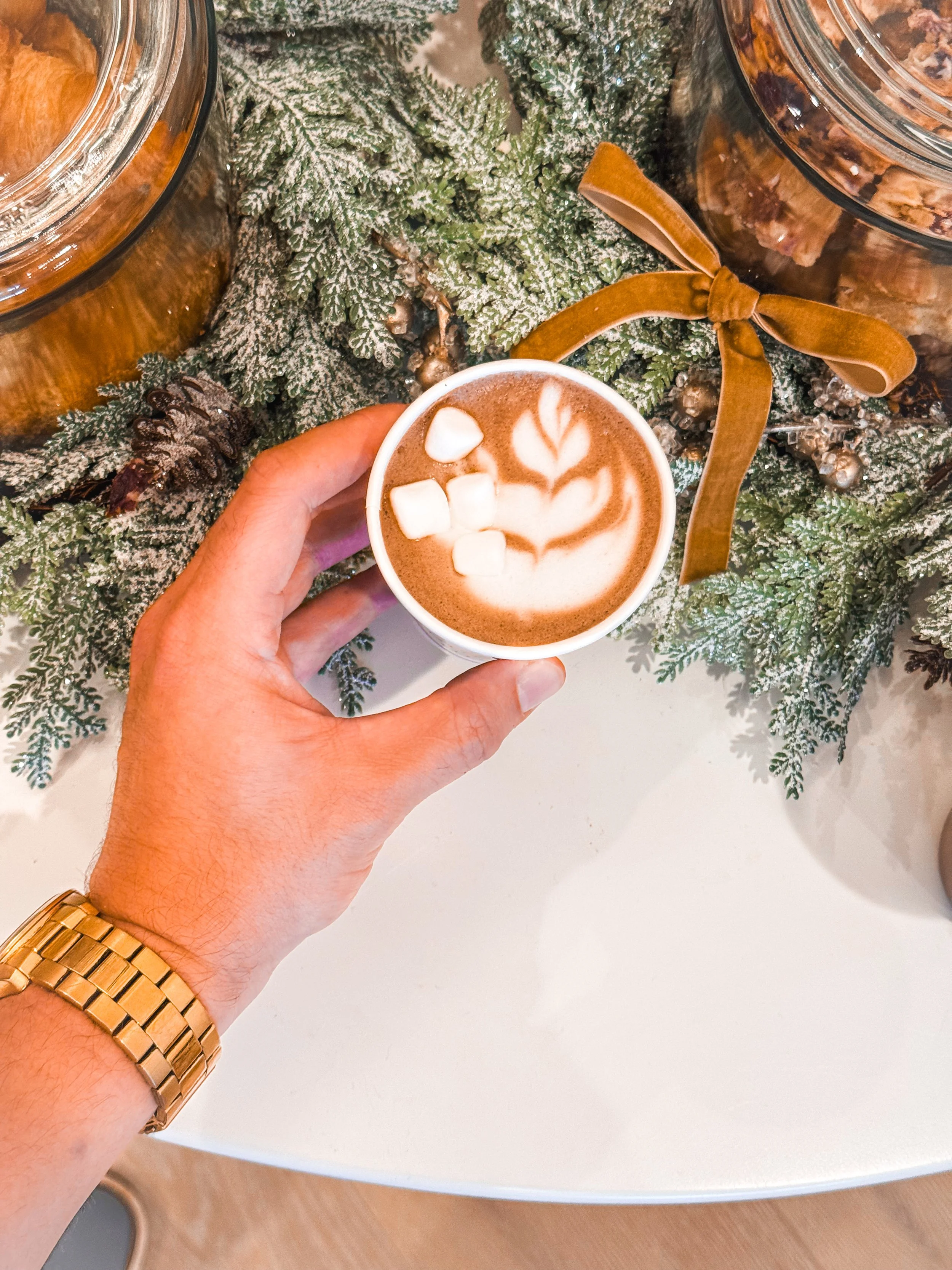 A hand holding a cup of hot chocolate by Bendecido Coffee with marshmallows and latte art, in front of Christmas greenery and jars of cookies on a white table.