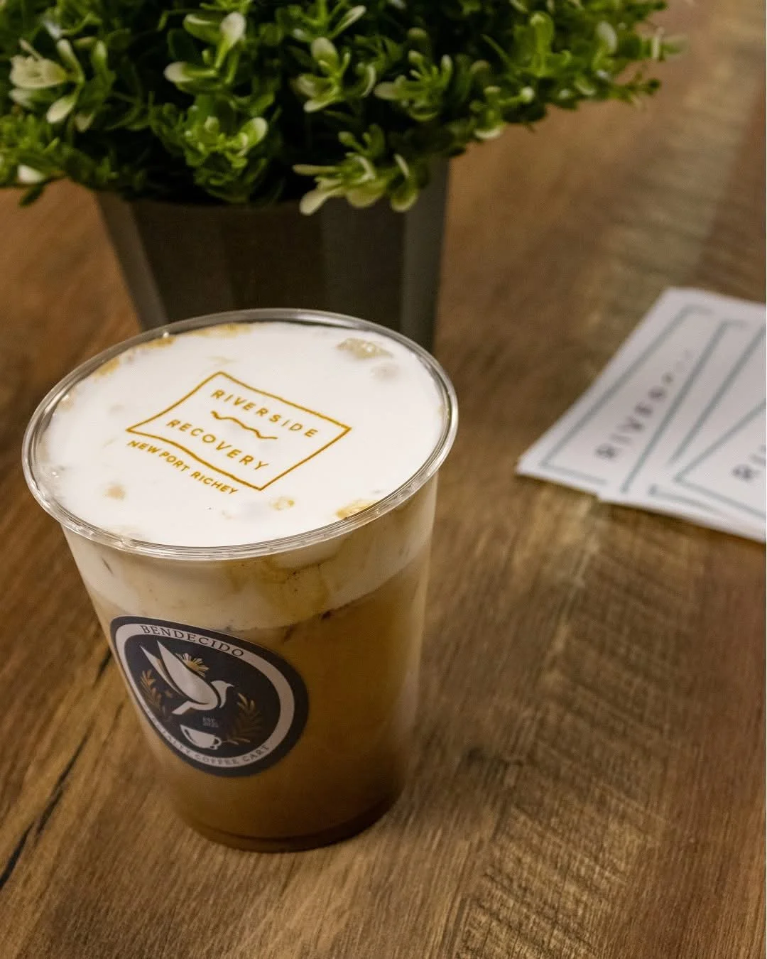 A plastic cup of iced coffee with foam and a Riverside Recovery sticker, placed on a wooden table near a green potted plant and some receipts.