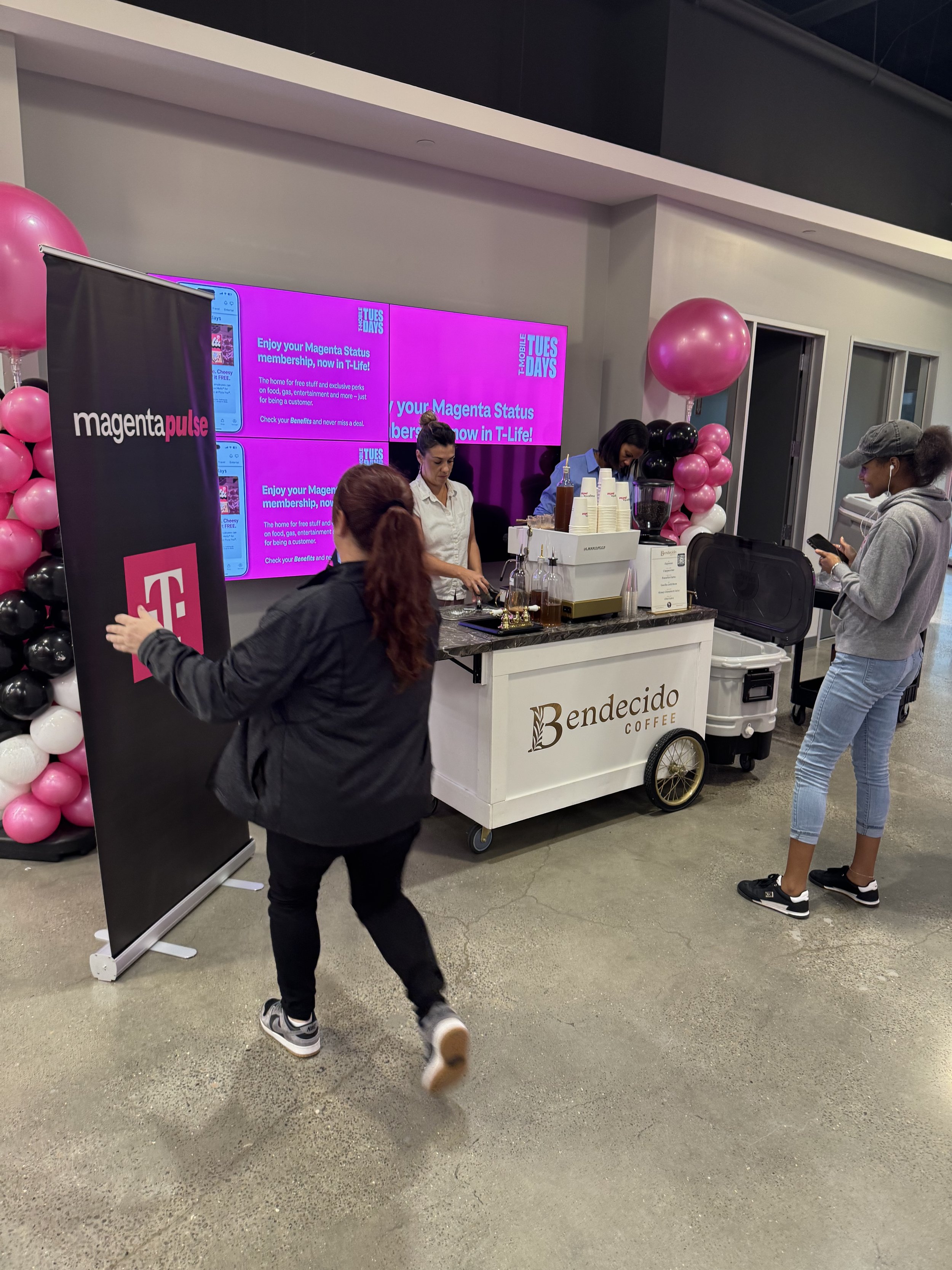 A coffee cart labeled 'Bendecido Coffee' at an event with pink and black balloons nearby. A woman is standing at the counter, while another woman looks at her phone. A large screen in the background displays an advertisement for Magenta T-Mobile with