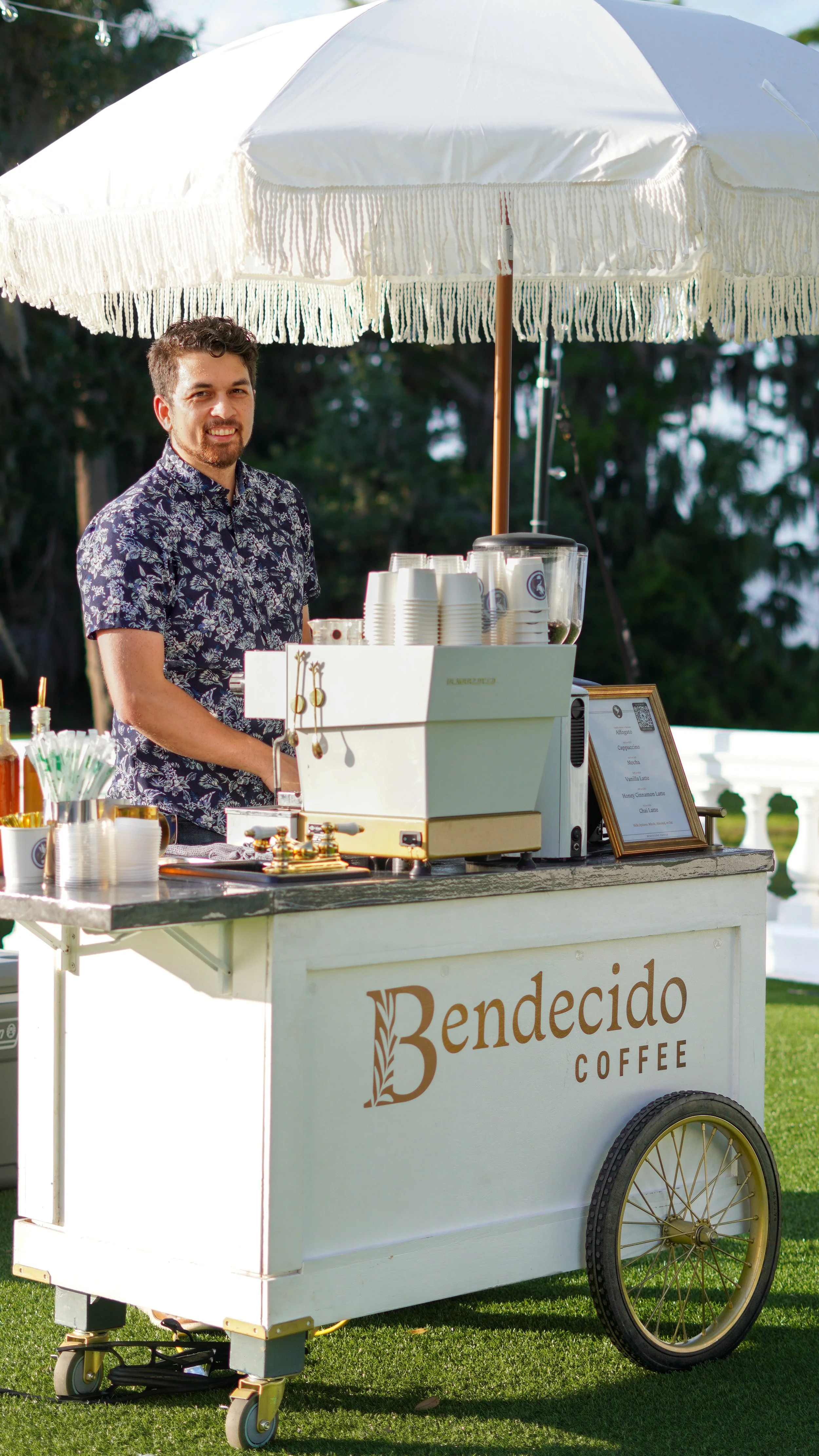 Smiling man standing behind a white coffee cart labeled "Bendecito Coffee" with a large white parasol on a sunny outdoor setting.