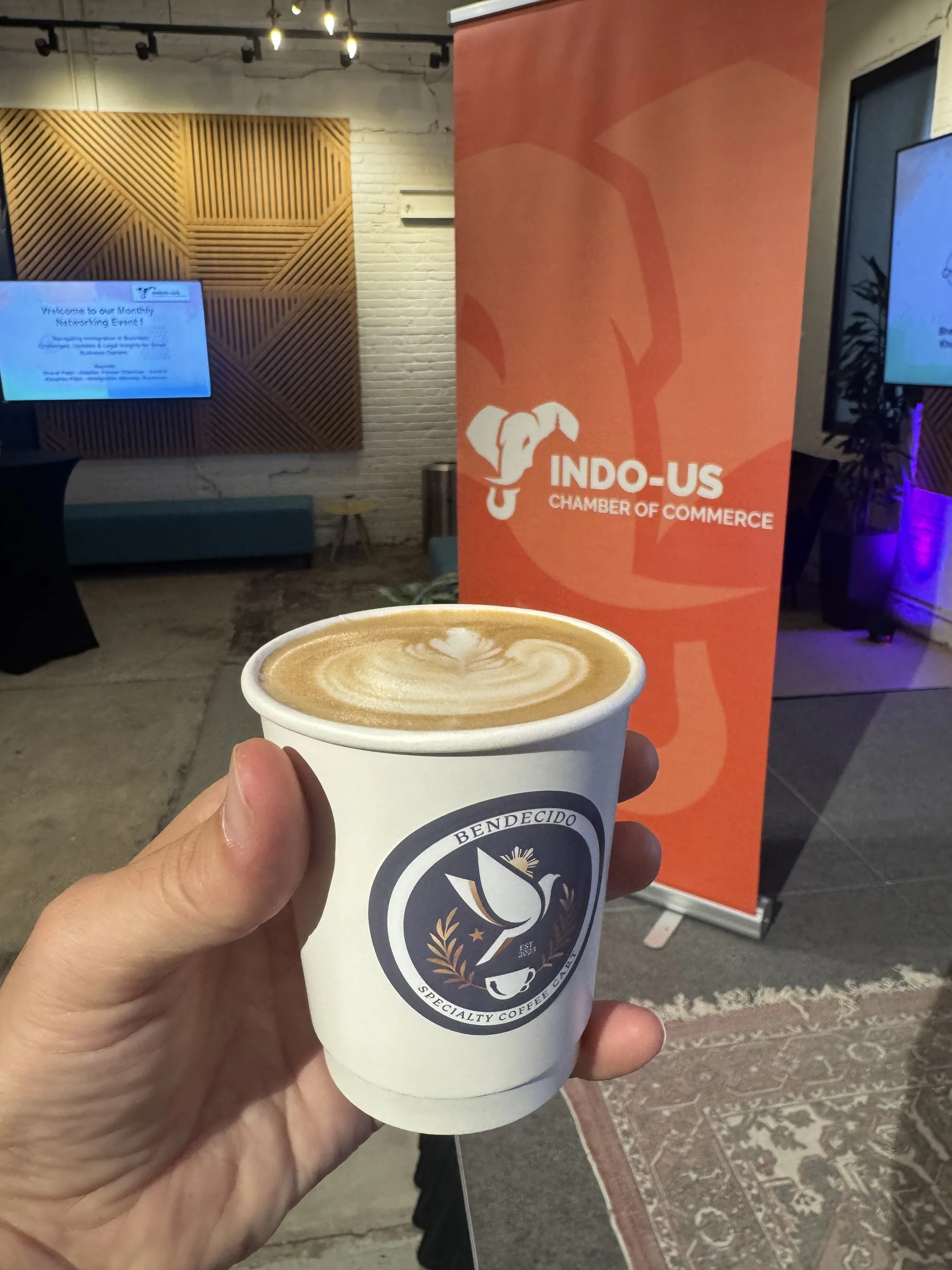 A hand holding a cup of coffee with latte art, in front of an Indo-US Chamber of Commerce banner at a networking event.