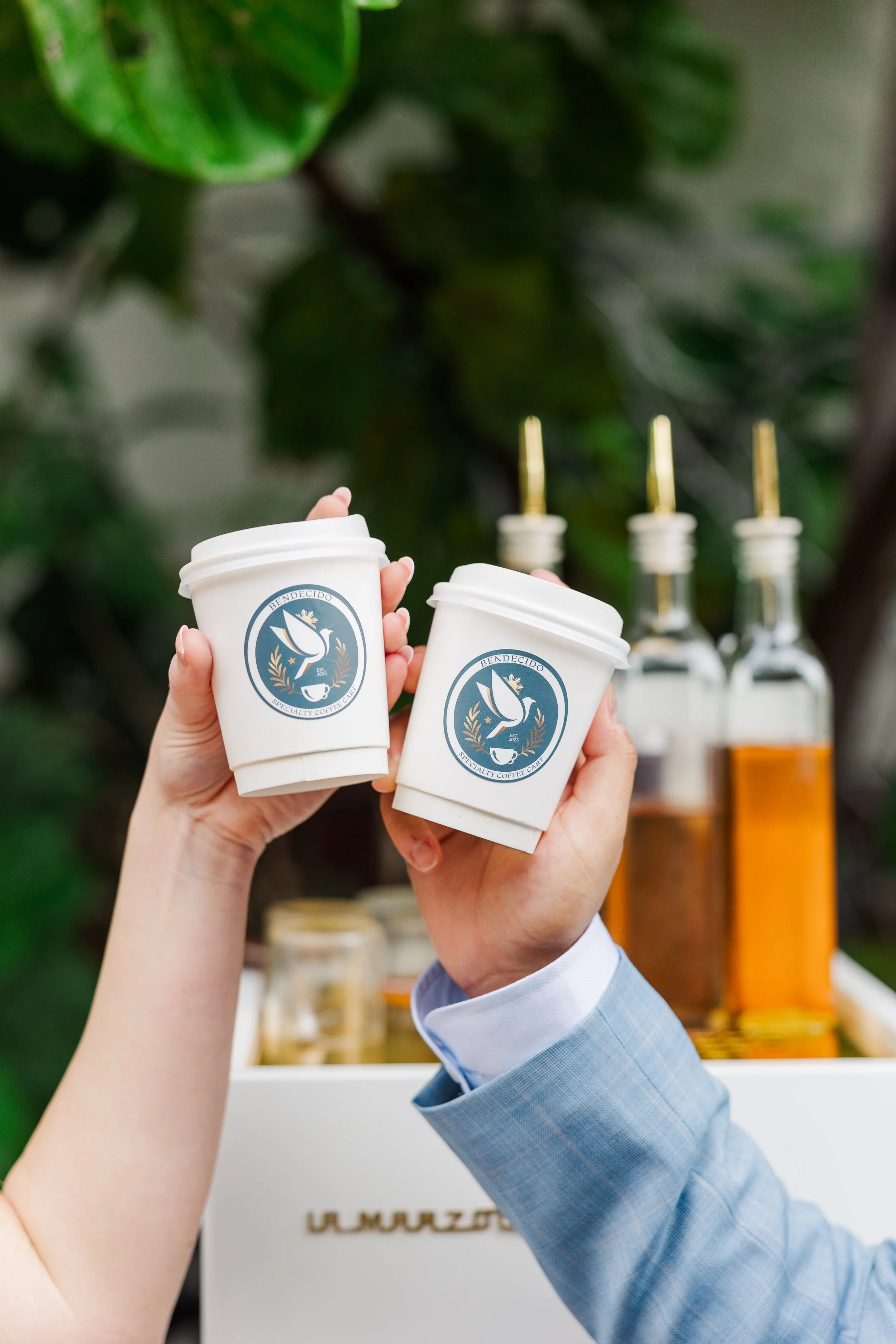 Two hands holding white coffee cups with a logo that has a dove and a laurel, in front of bottles of syrup with golden pour spouts.