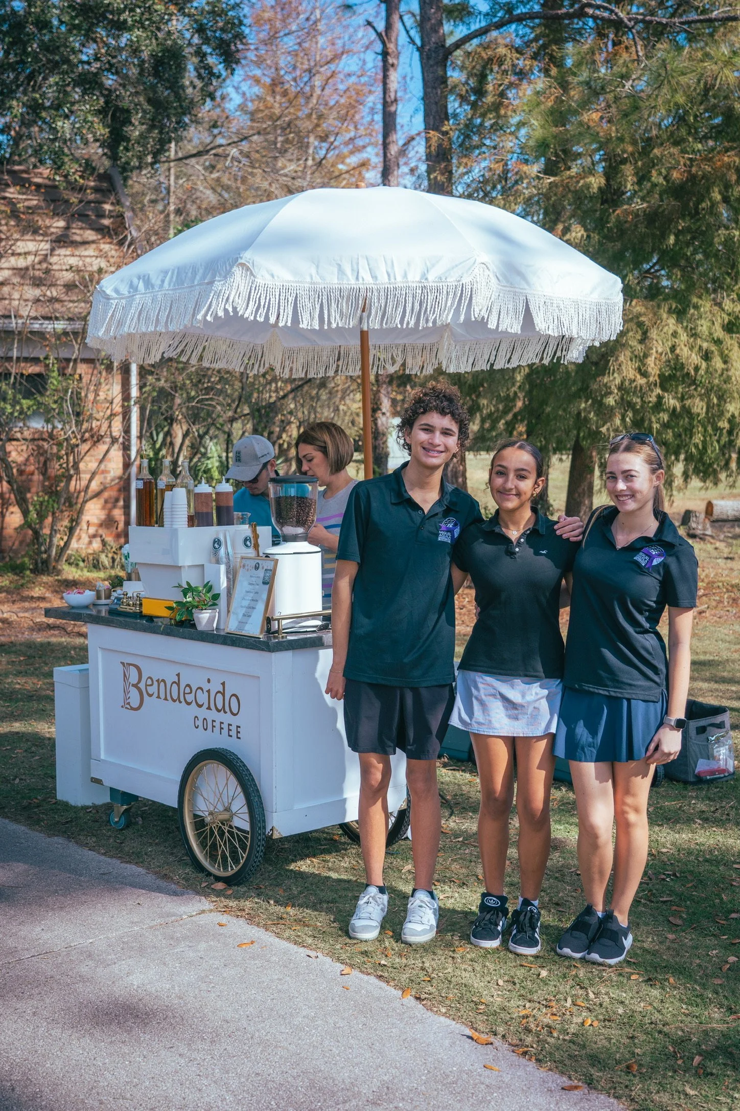 Three smiling Bendecido Coffee staff in navy polos beside a cart under a white umbrella