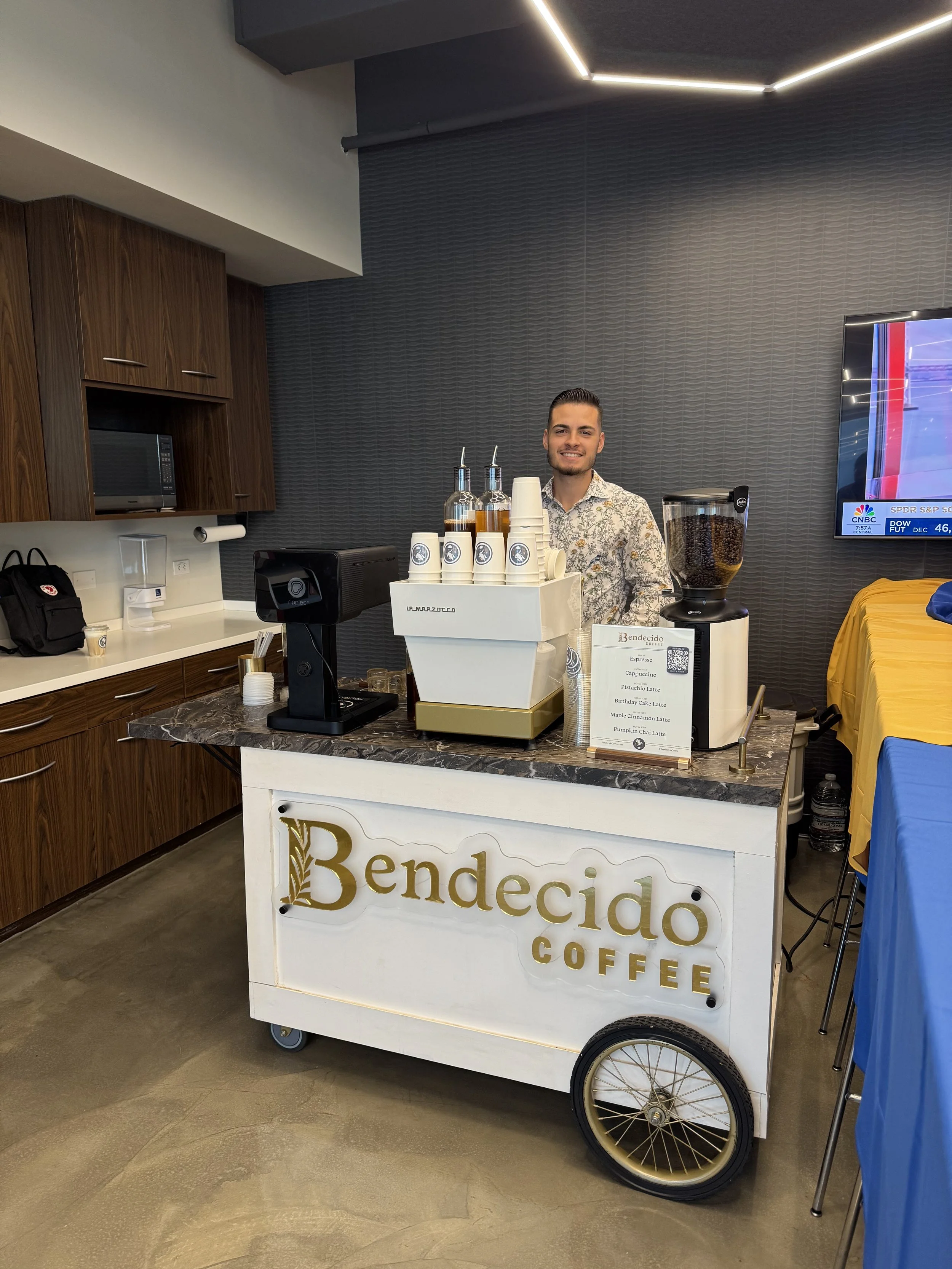 A man standing behind a coffee cart with 'Bendecidō Coffee' written on it, serving coffee at an indoor event.