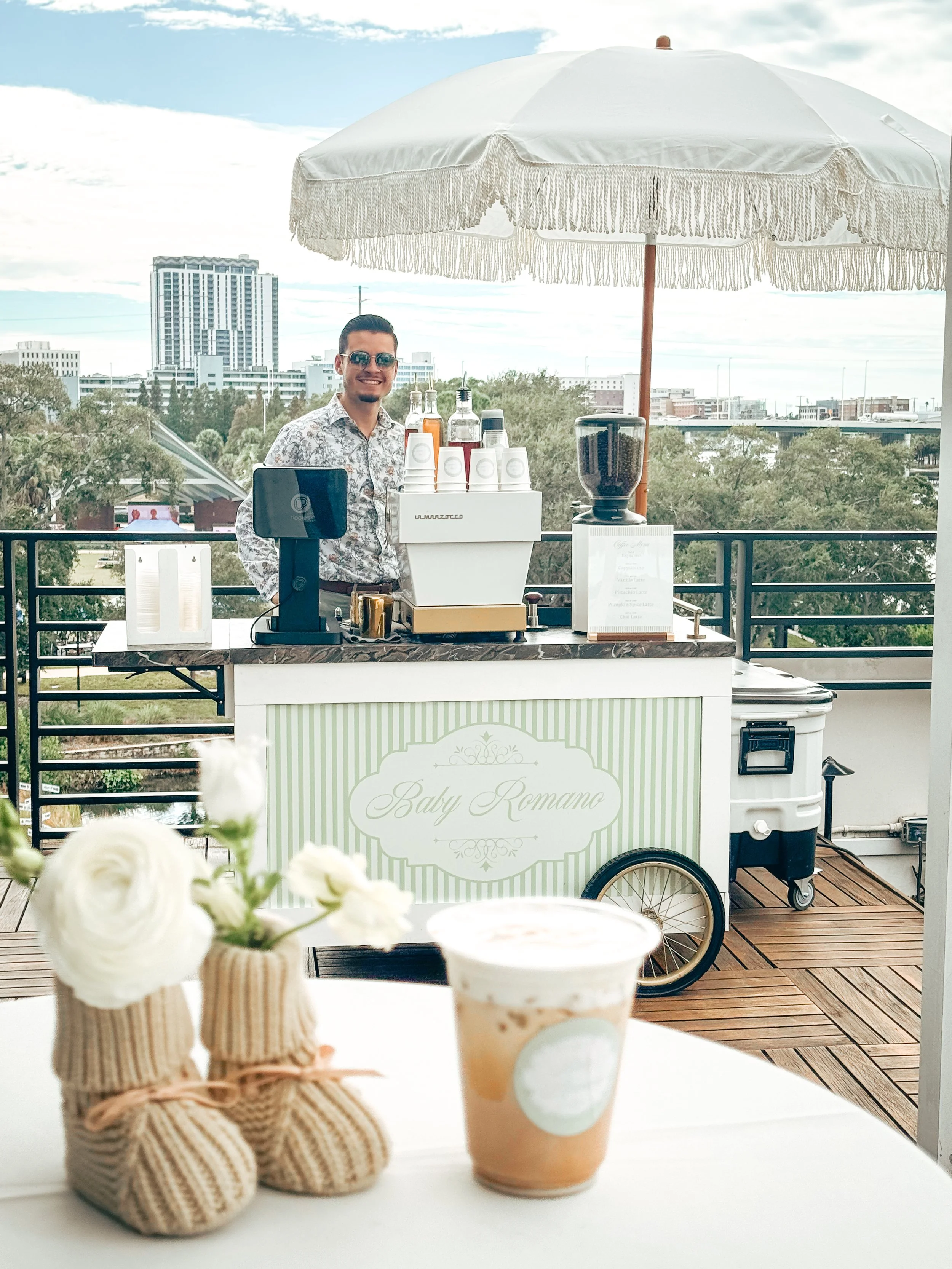 A happy man standing behind a baby-themed coffee cart with a sign that reads "Baby Romano". The cart has various bottles, a blender, and cups, with an umbrella providing shade. In the foreground, there are small knitted baby boots with flowers and an